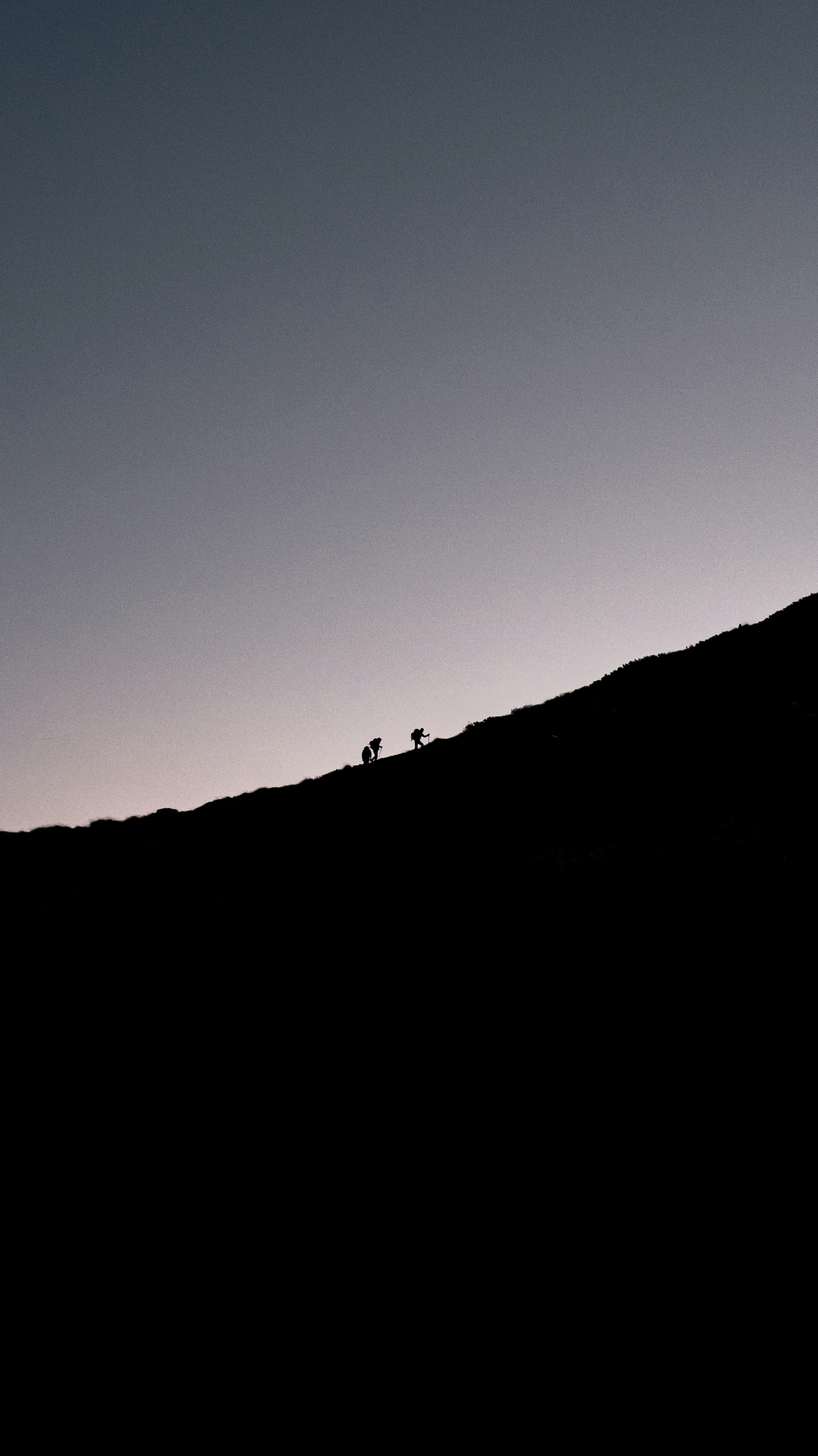 Two hikers ascend a dark mountain slope at dusk.