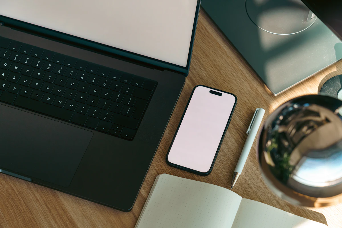Laptop terbuka dengan ponsel dan buku catatan tergeletak di samping keyboard di atas meja kayu
