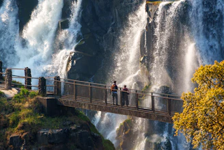 Two people on a bridge near a powerful waterfall
