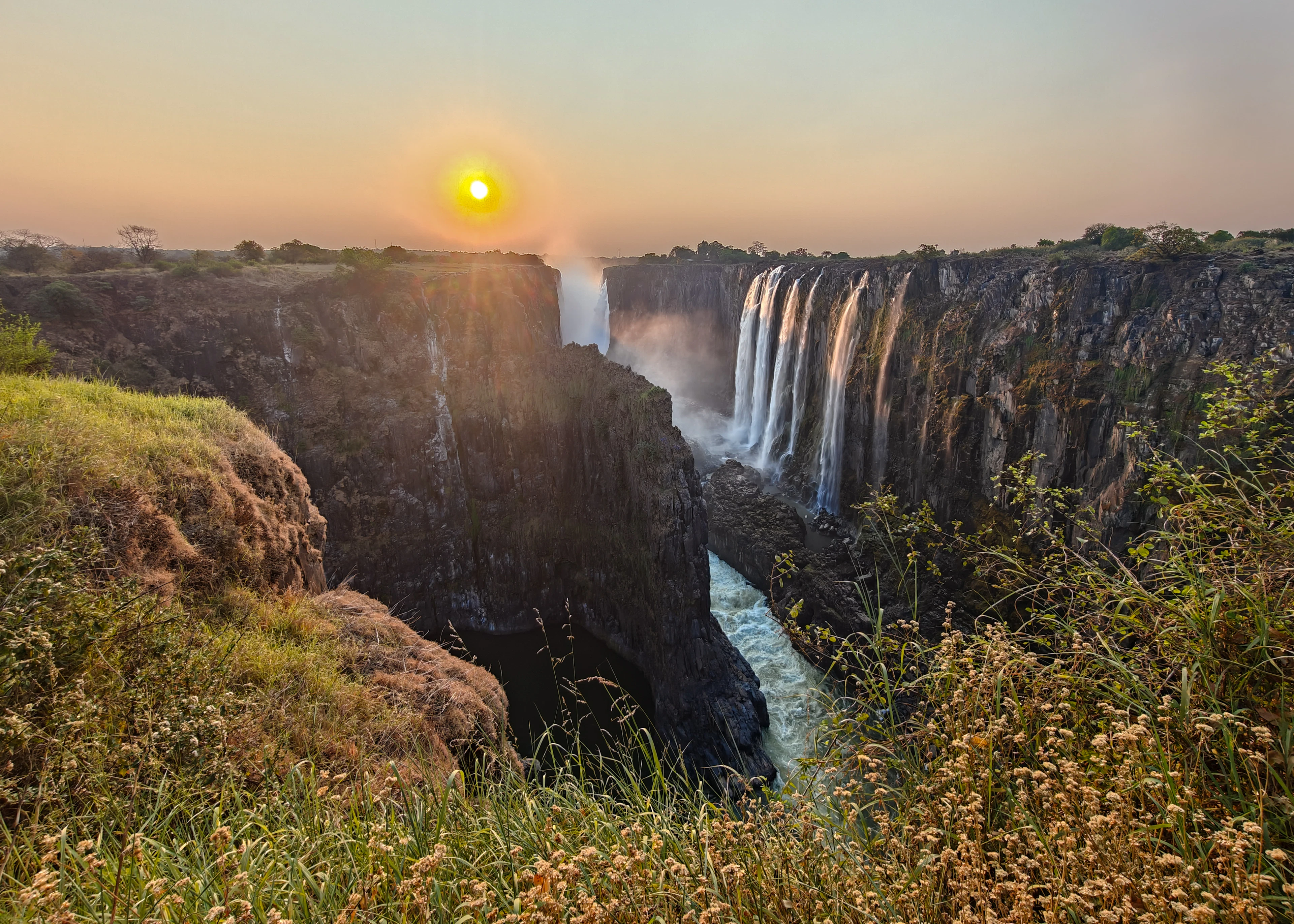 A wide dawn view of Victoria Falls cascading along the cliff line, sun rising above the gorge.