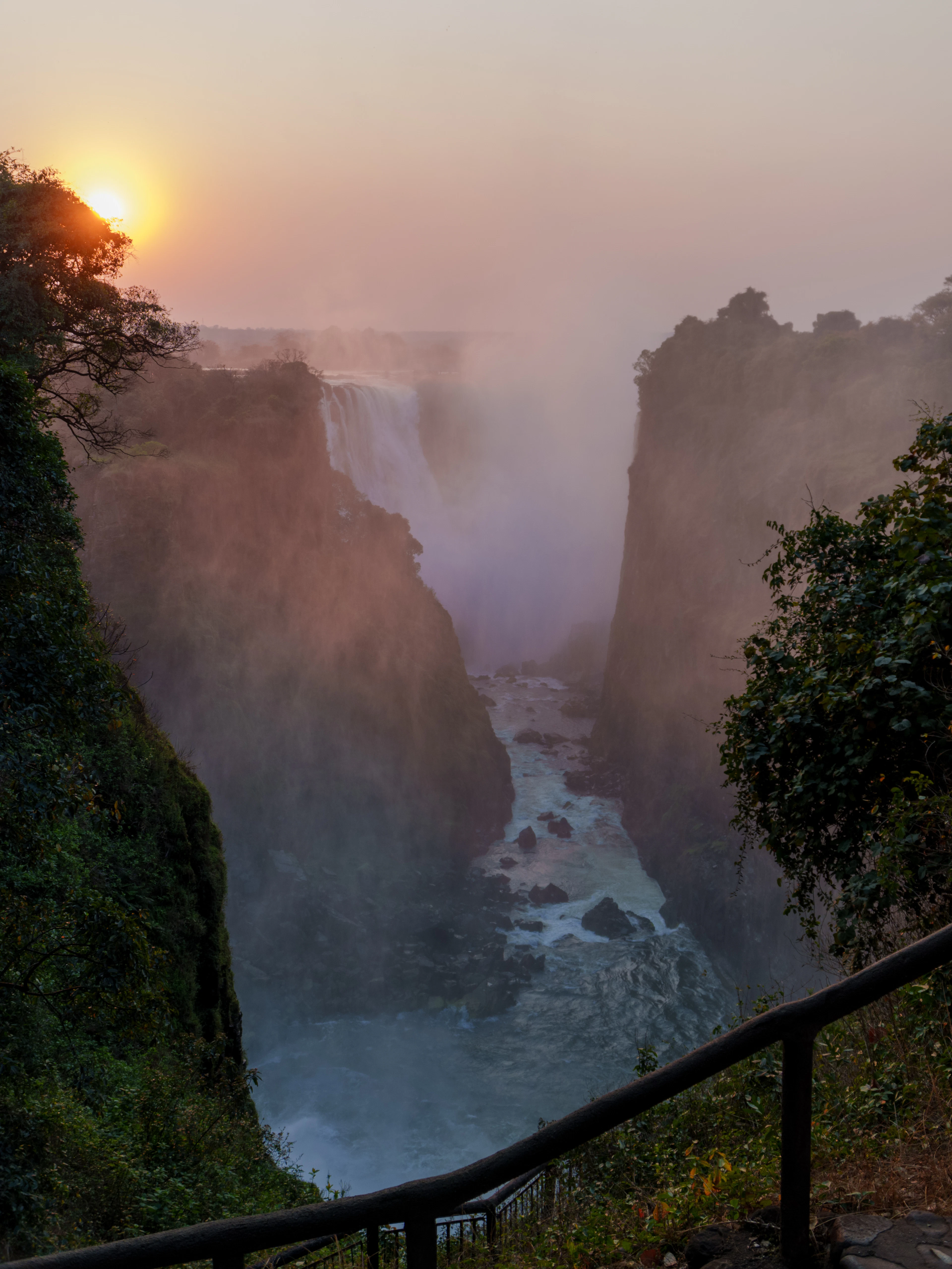Sunrise glows through mist rising off the Victoria Falls gorge, viewed from a platform railing.