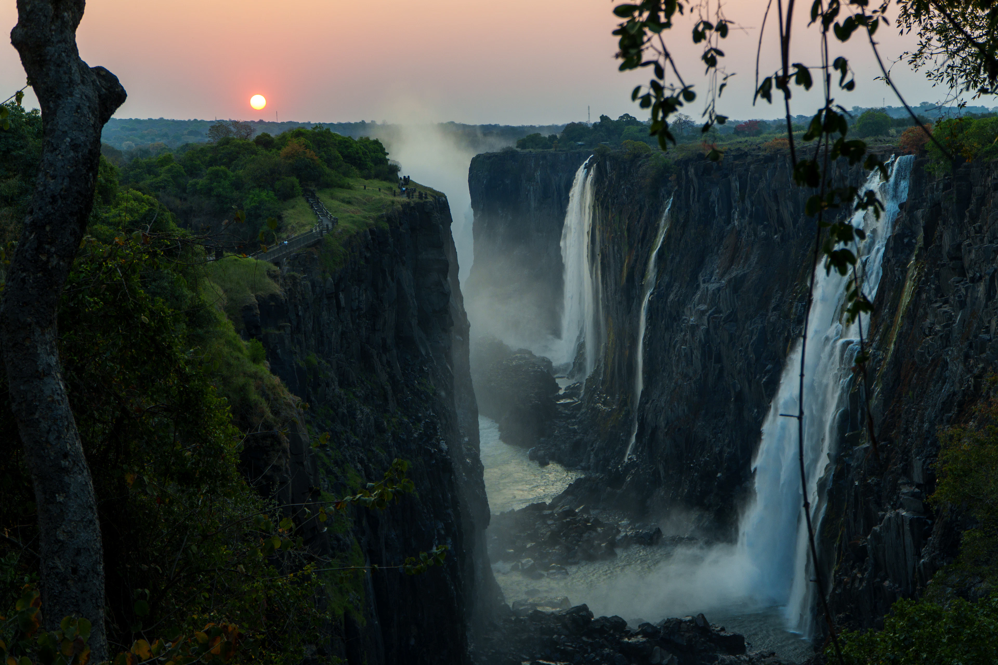 The sun low over Victoria Falls, curtains of water dropping into the narrow basalt gorge with spray rising from the chasm.