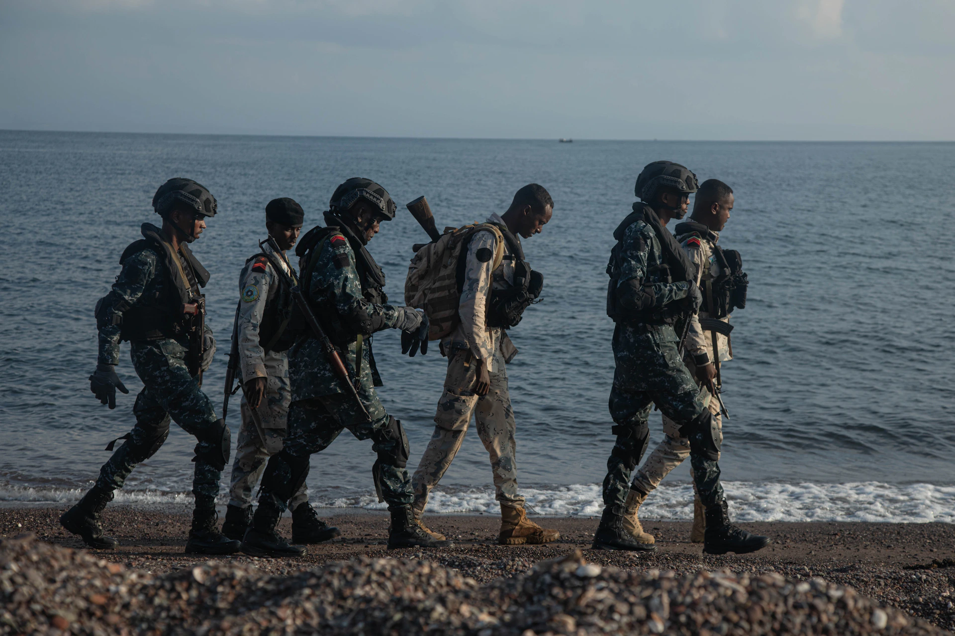 Soldiers walk along a rocky beach by the ocean.