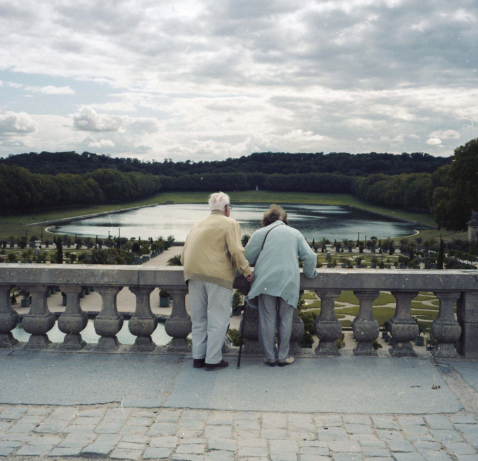 An older couple standing together above a formal garden and tree-lined grounds.