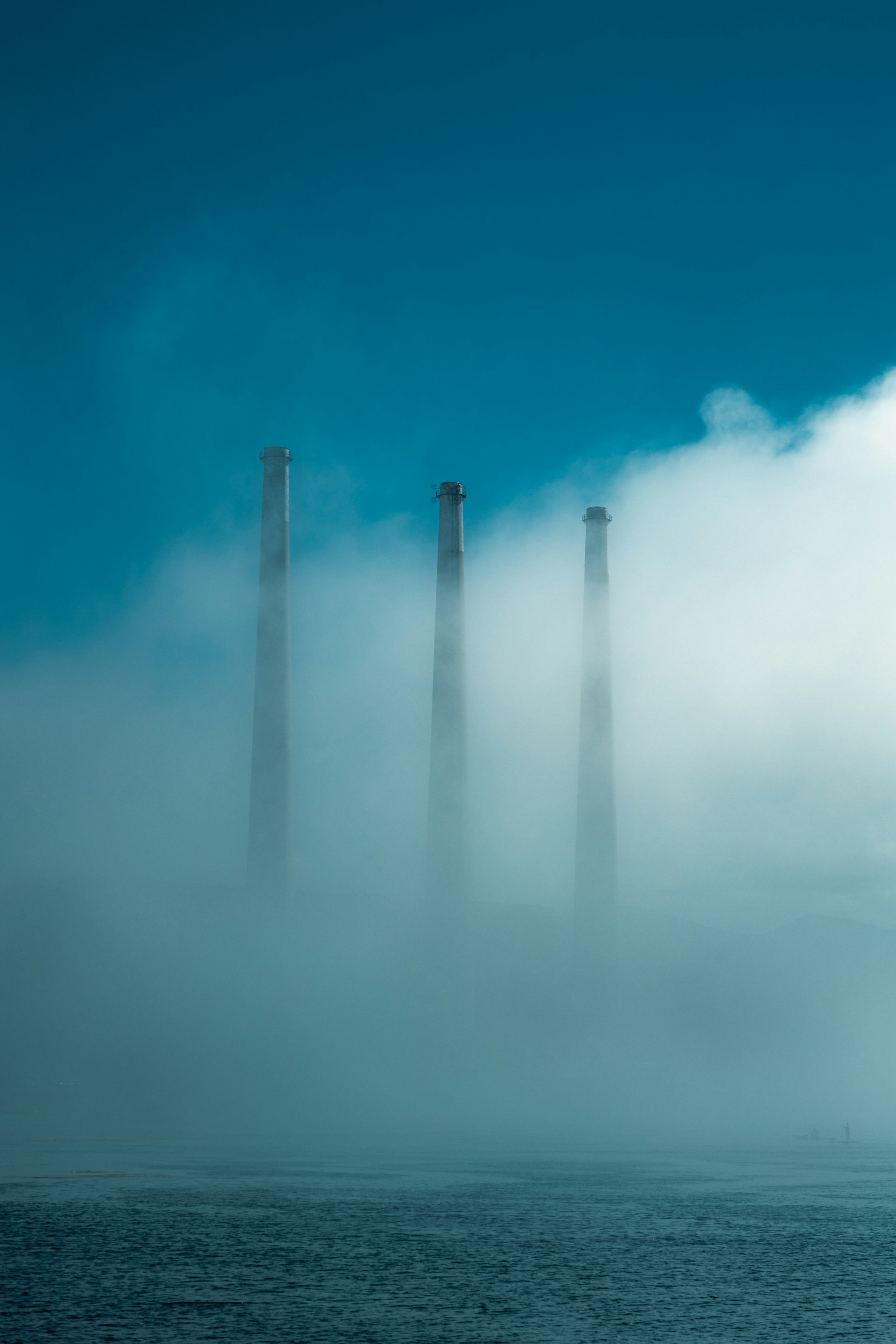 Three smokestacks emerge from thick fog over water.