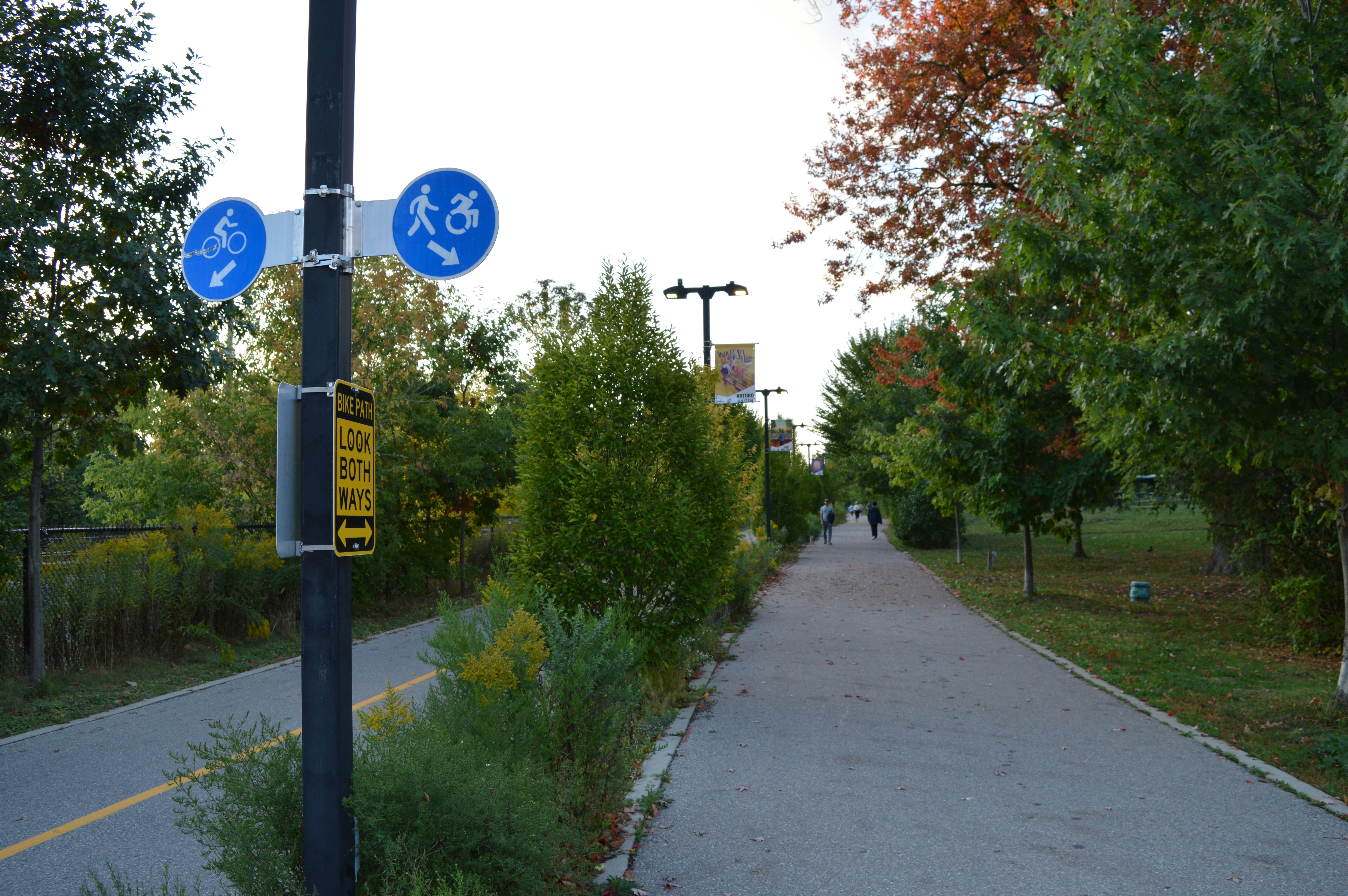 ทางเดินปูพื้นในสวนสาธารณะที่มีต้นไม้และป้าย