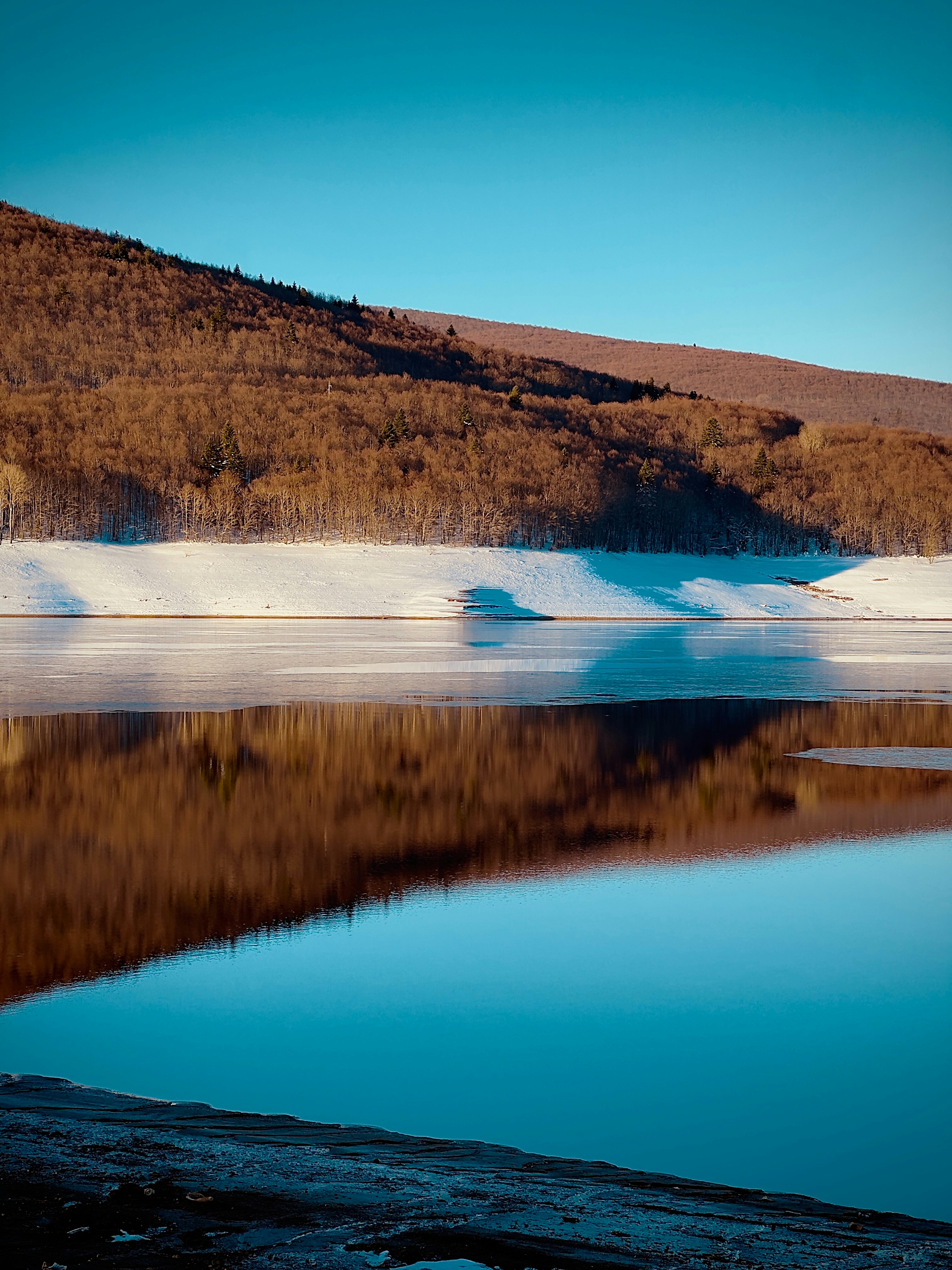 Calm lake reflecting brown hillside under clear sky.