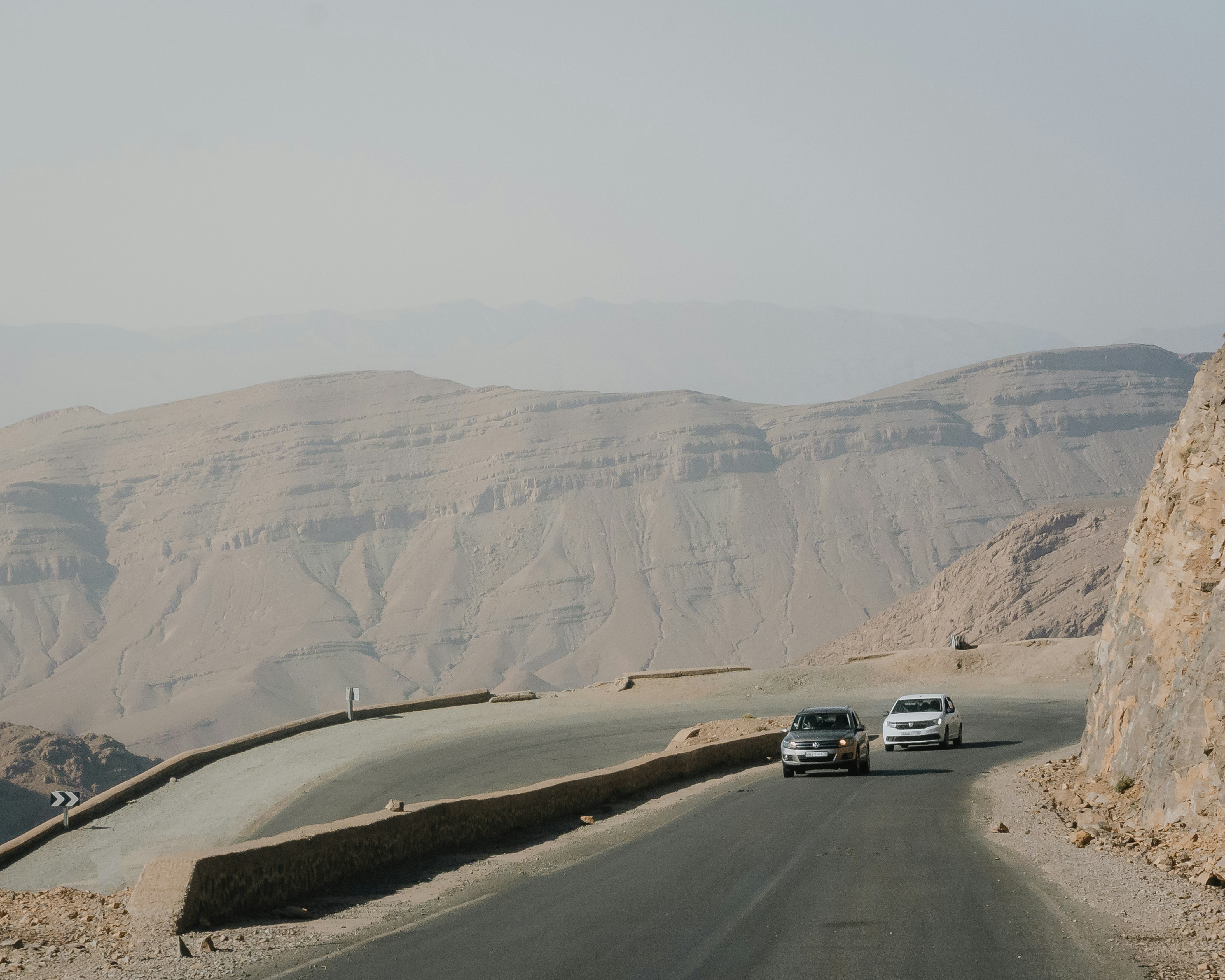Two cars drive on a winding road through desert mountains.