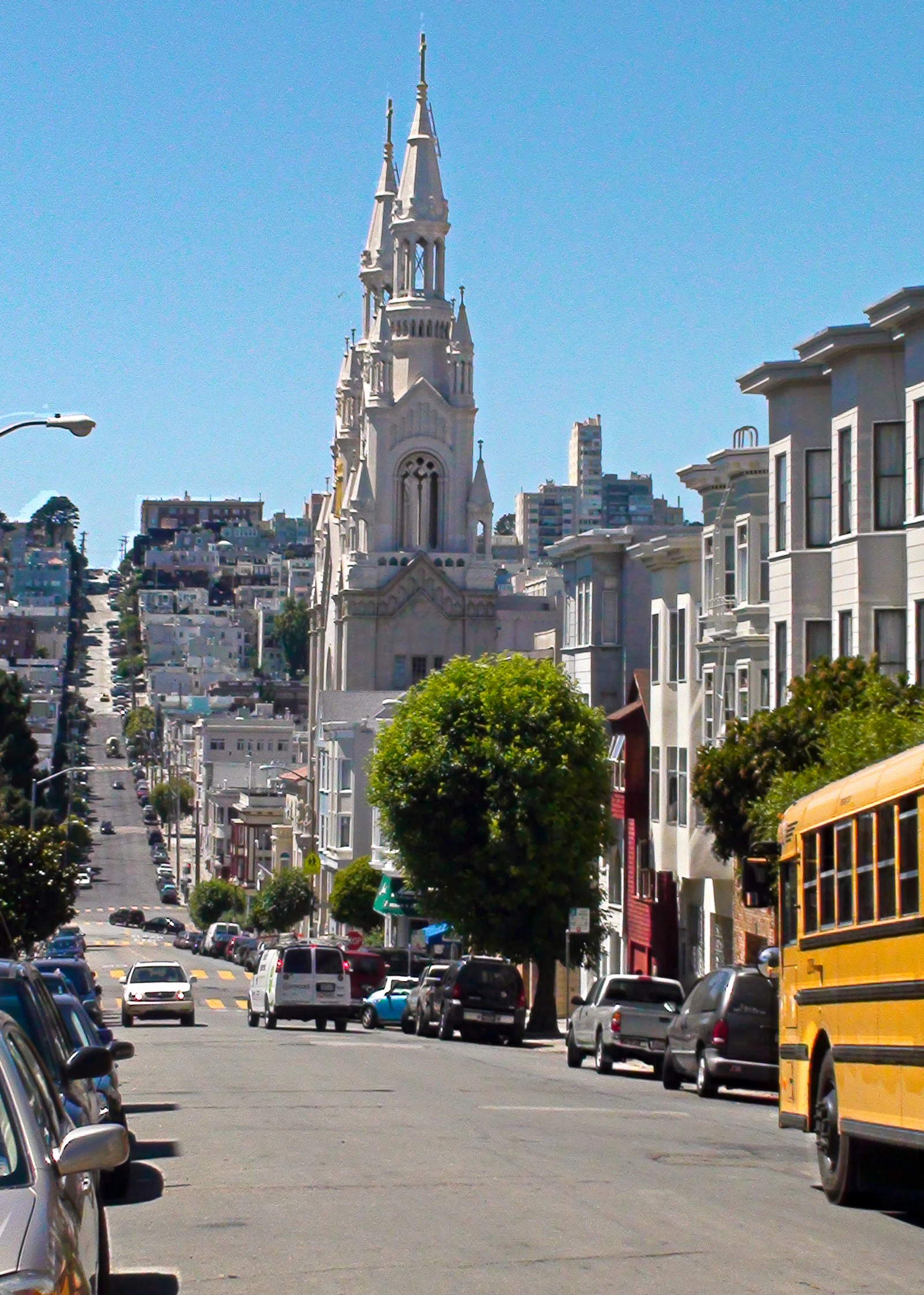 A yellow school bus parked on a steep San Francisco street, with Saints Peter and Paul Church in the background