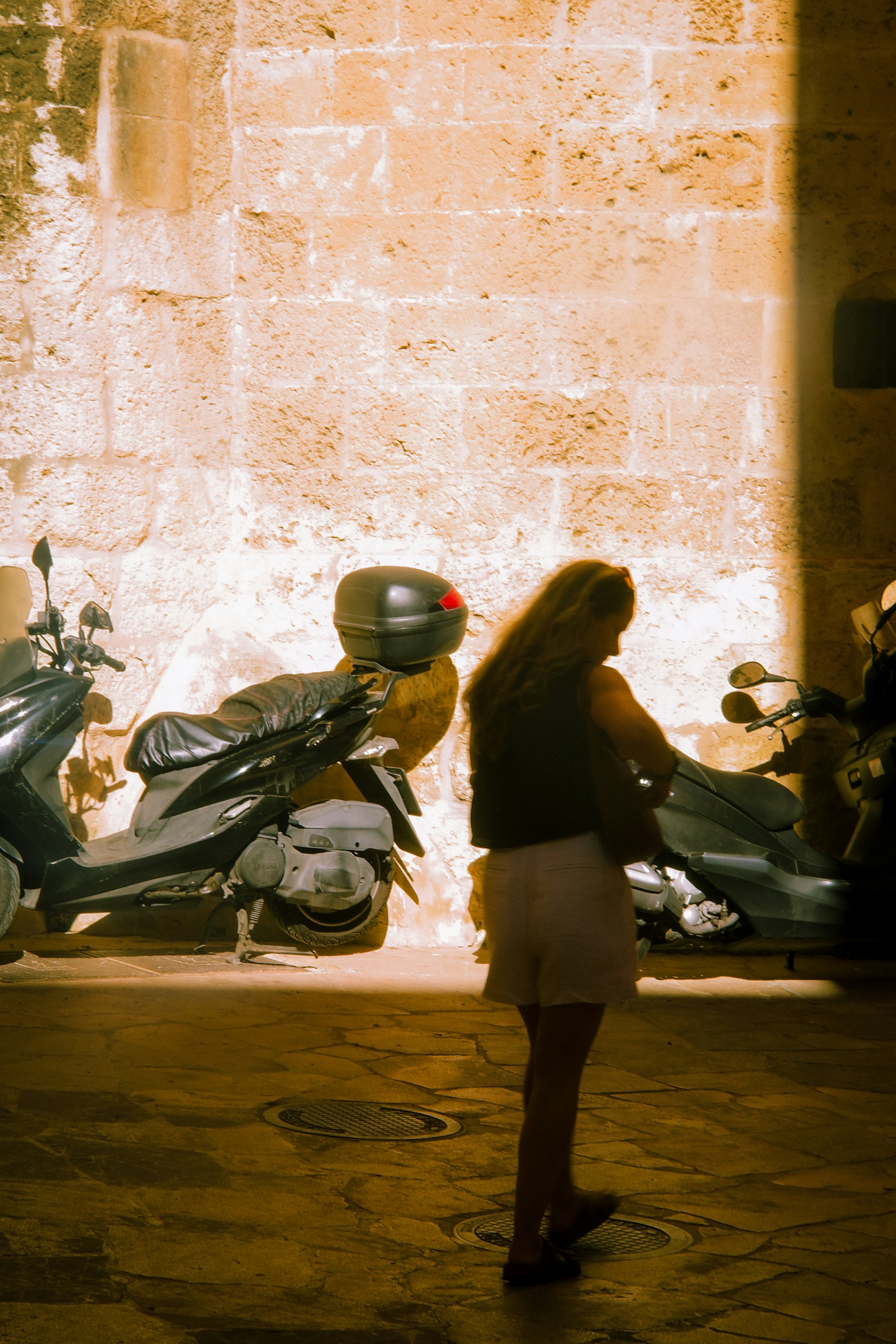 Woman standing near parked scooters by stone wall.