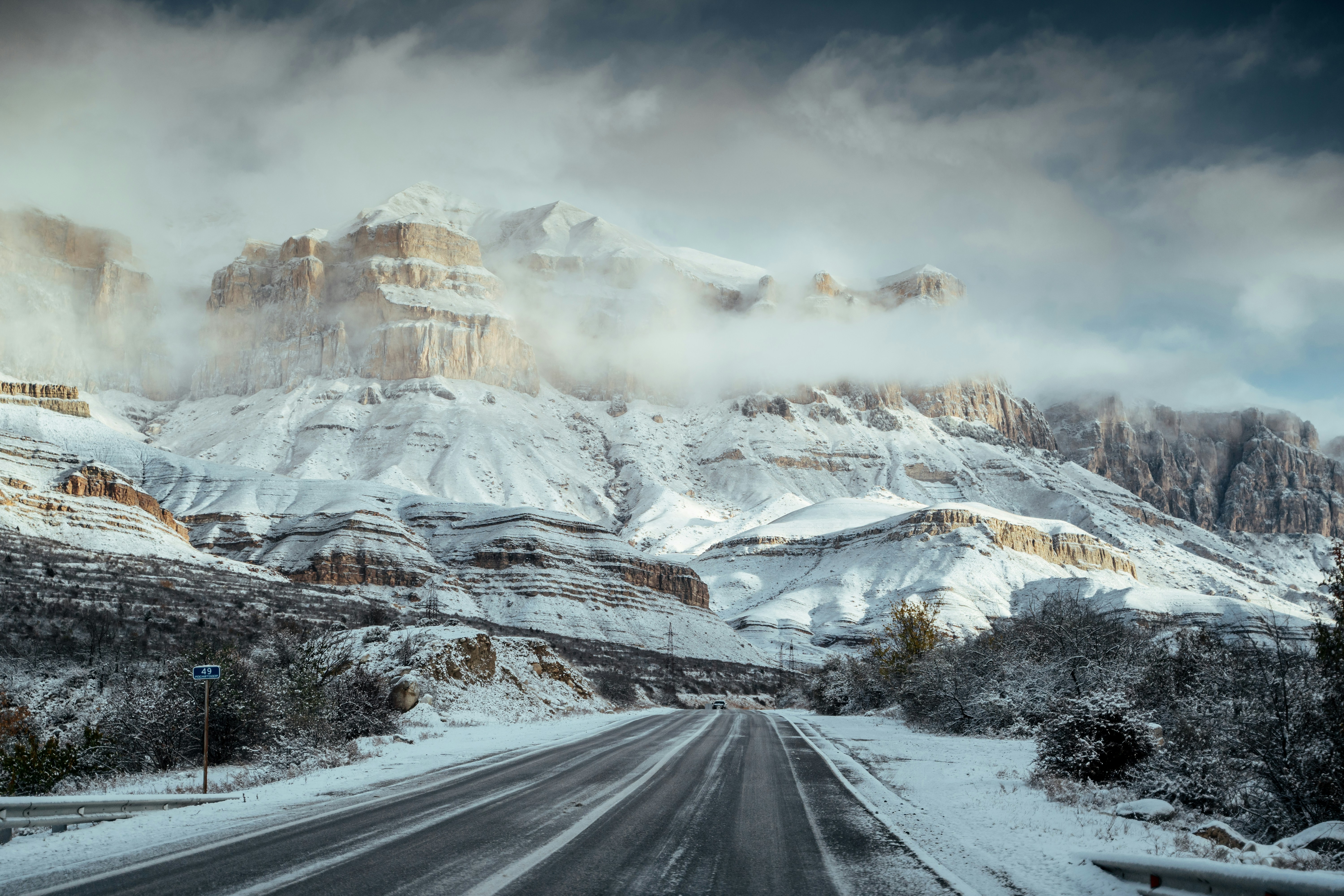 Snowy mountain road with clouds and rugged peaks.