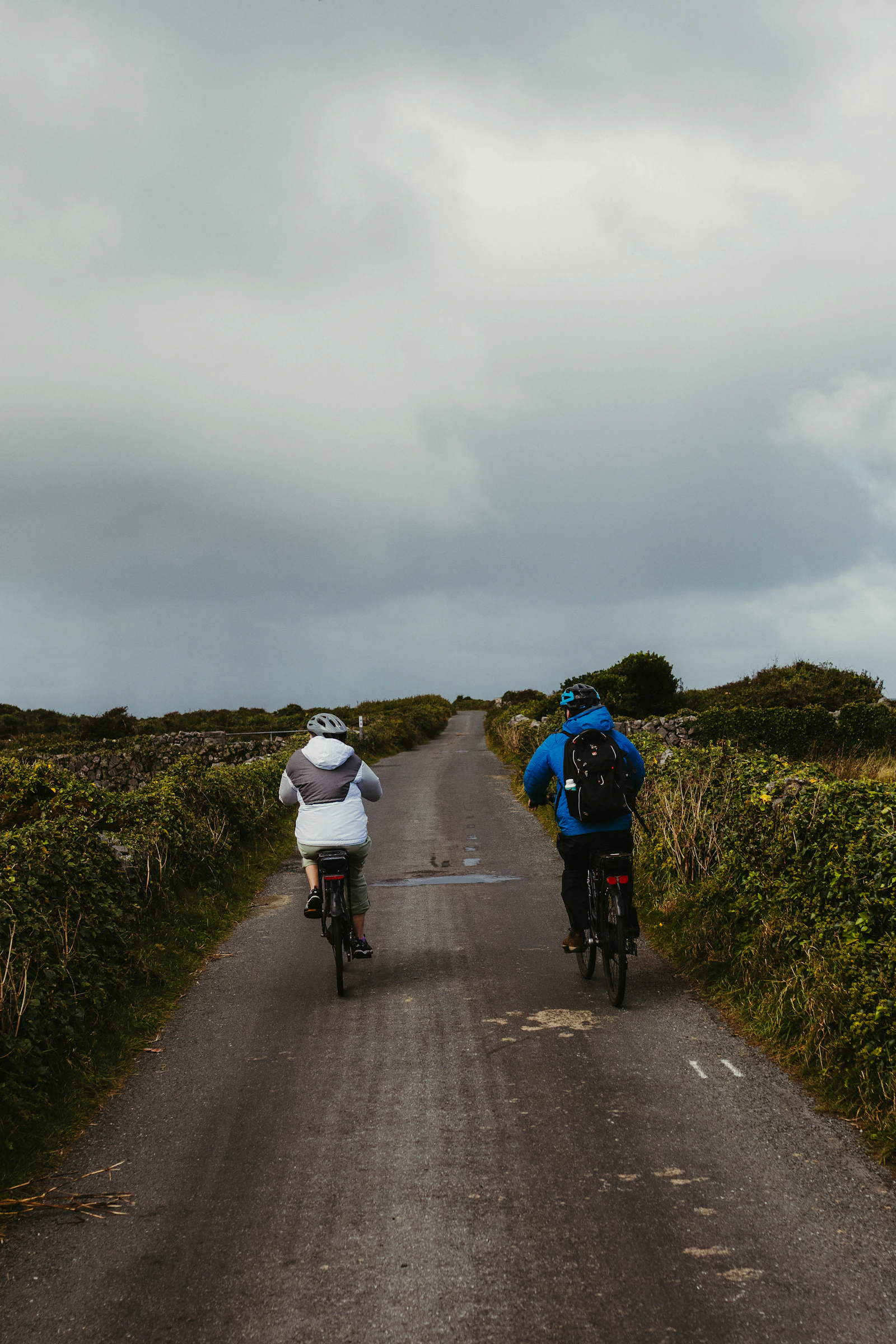 Two cyclists in jackets ride away up a narrow lane walled by hedges under heavy cloud