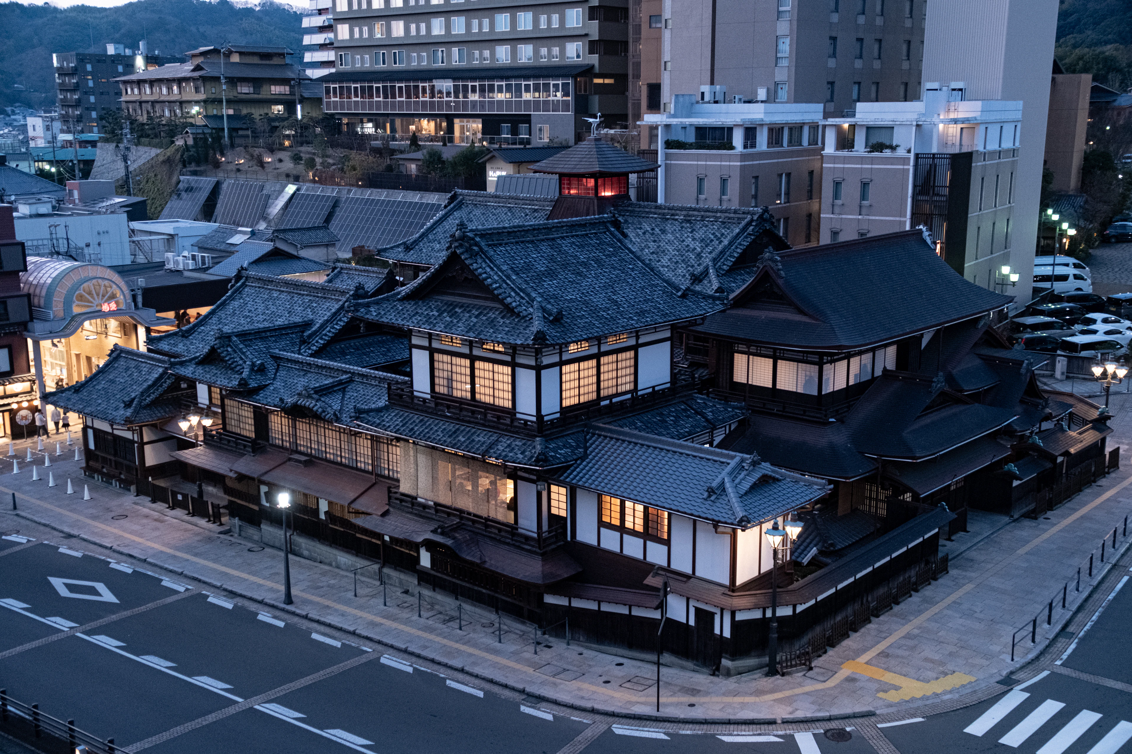 An old, many-roofed wooden bathhouse lit at dusk in a hot-spring quarter