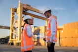 Two workers in hard hats discuss plans near shipping containers.