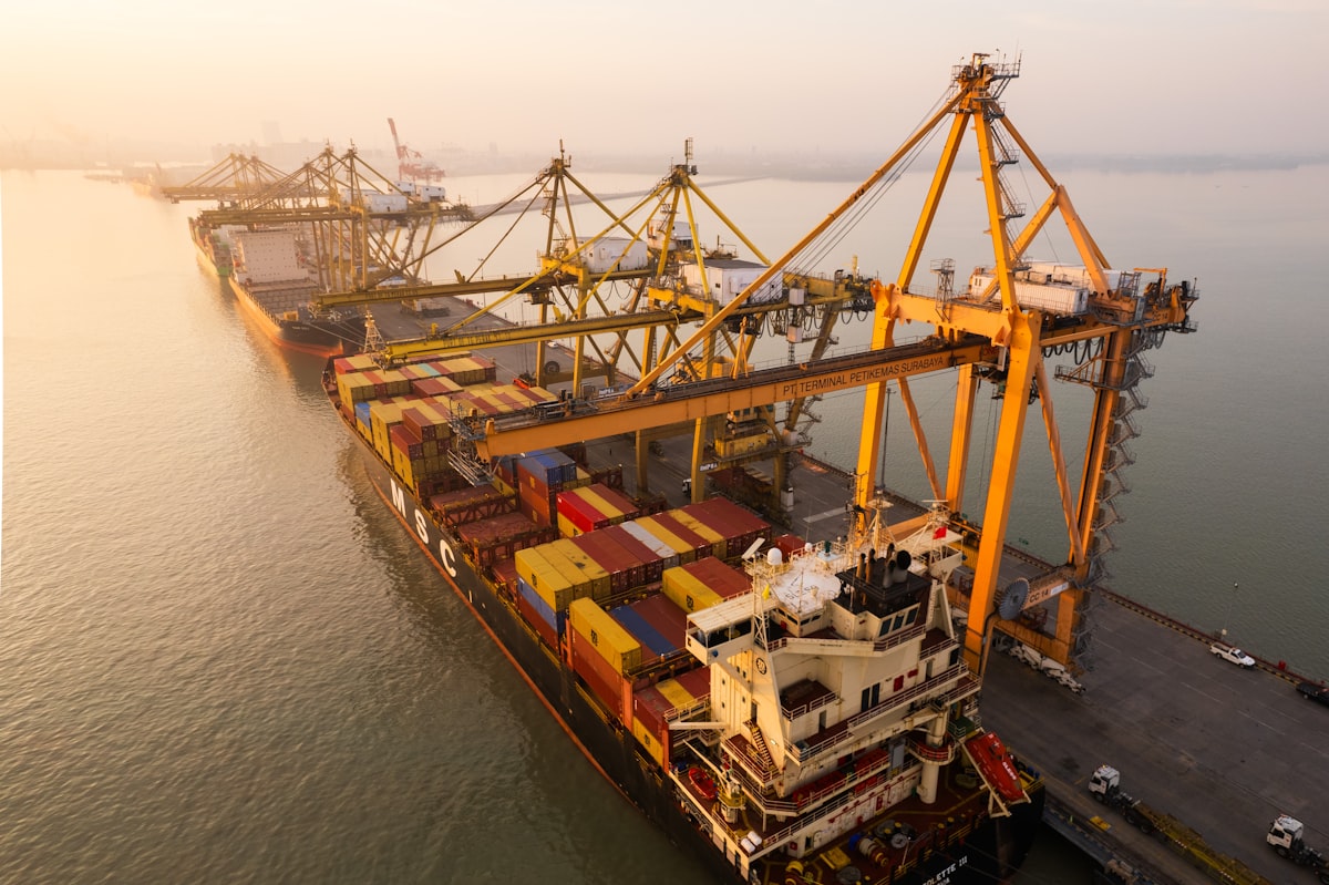 Aerial view of a cargo ship being loaded with shipping containers at a busy commercial port with large yellow cranes
