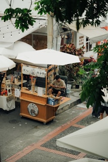 Man sits at a street food cart under umbrellas.