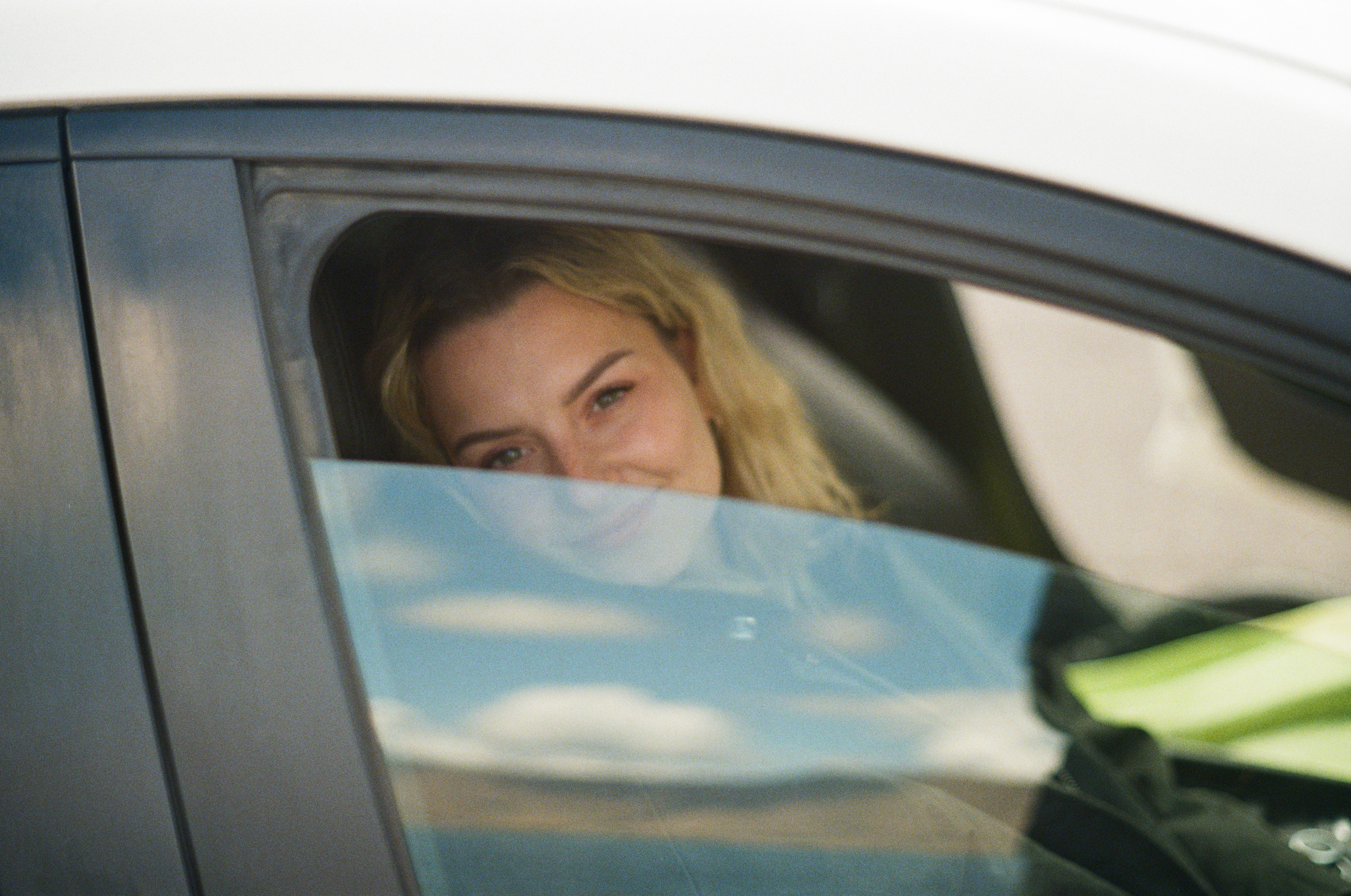Young woman smiling from the driver's seat of a car, with reflections of clouds visible on the window. A candid capture of a fleeting moment.