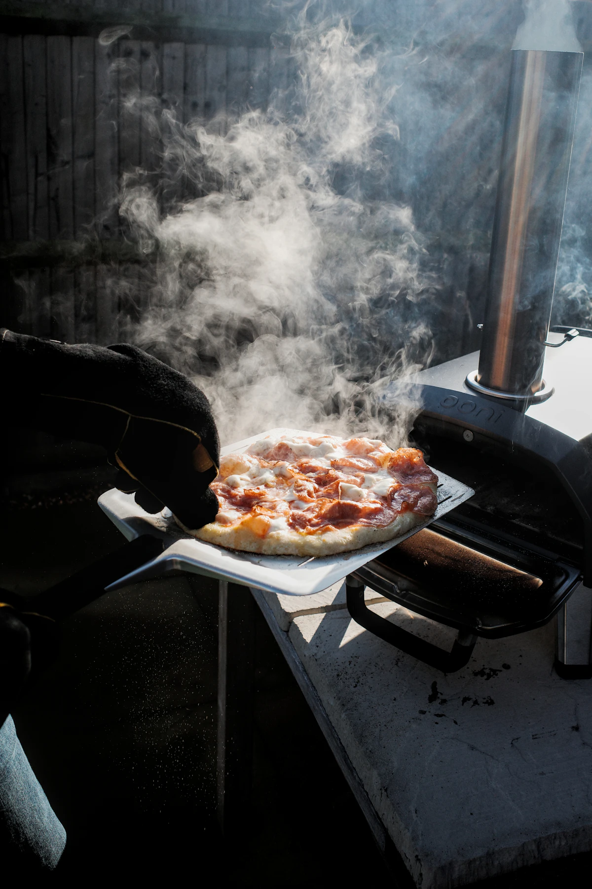 A finished pizza lifted out of a wood-fired oven on a steel peel, steam coming off it