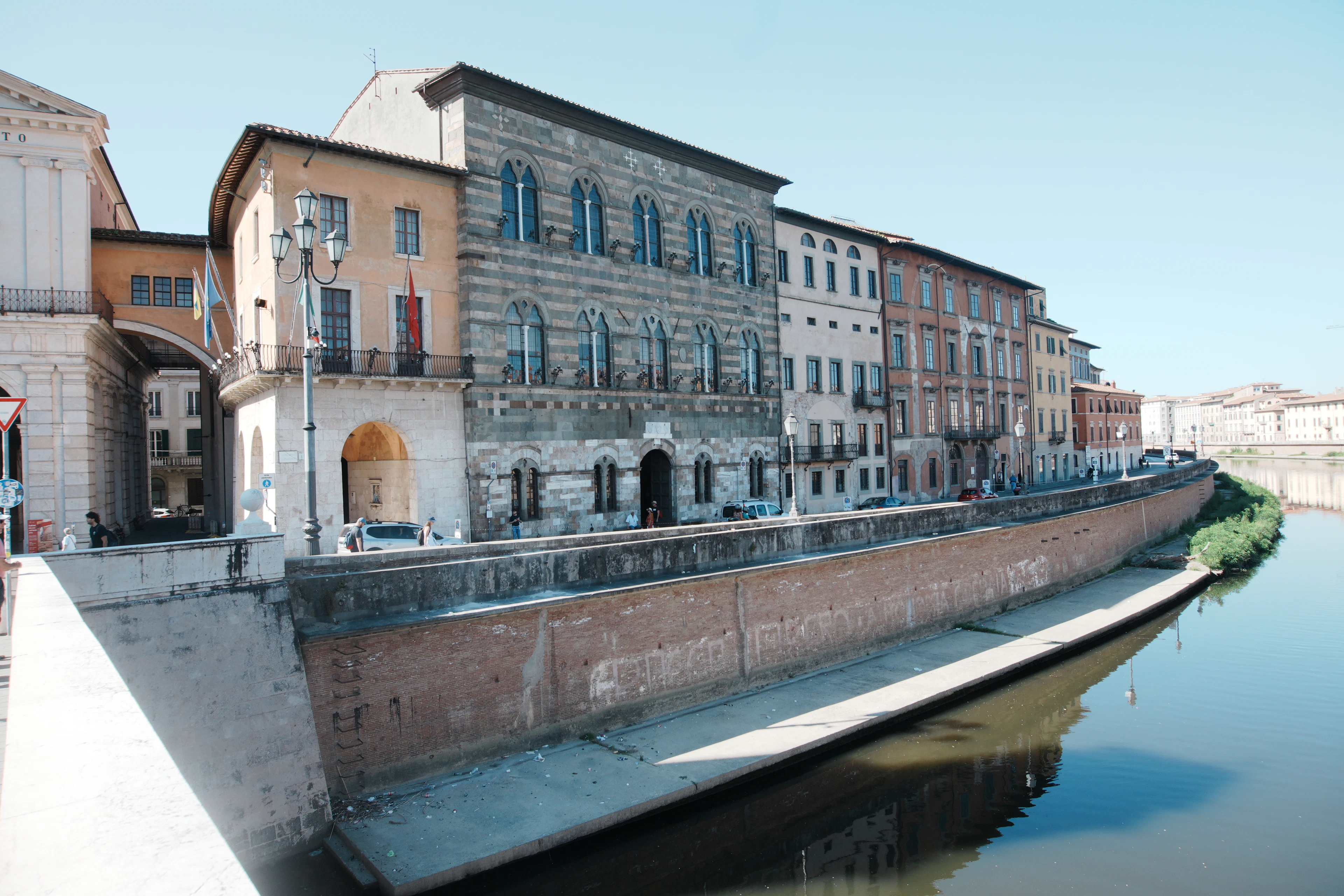Historic buildings lining the Arno river embankment in Pisa under a clear blue sky.