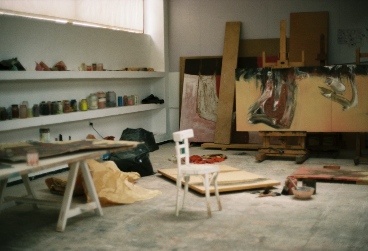 A large unfinished canvas on the studio wall, shelves of paint beside it
