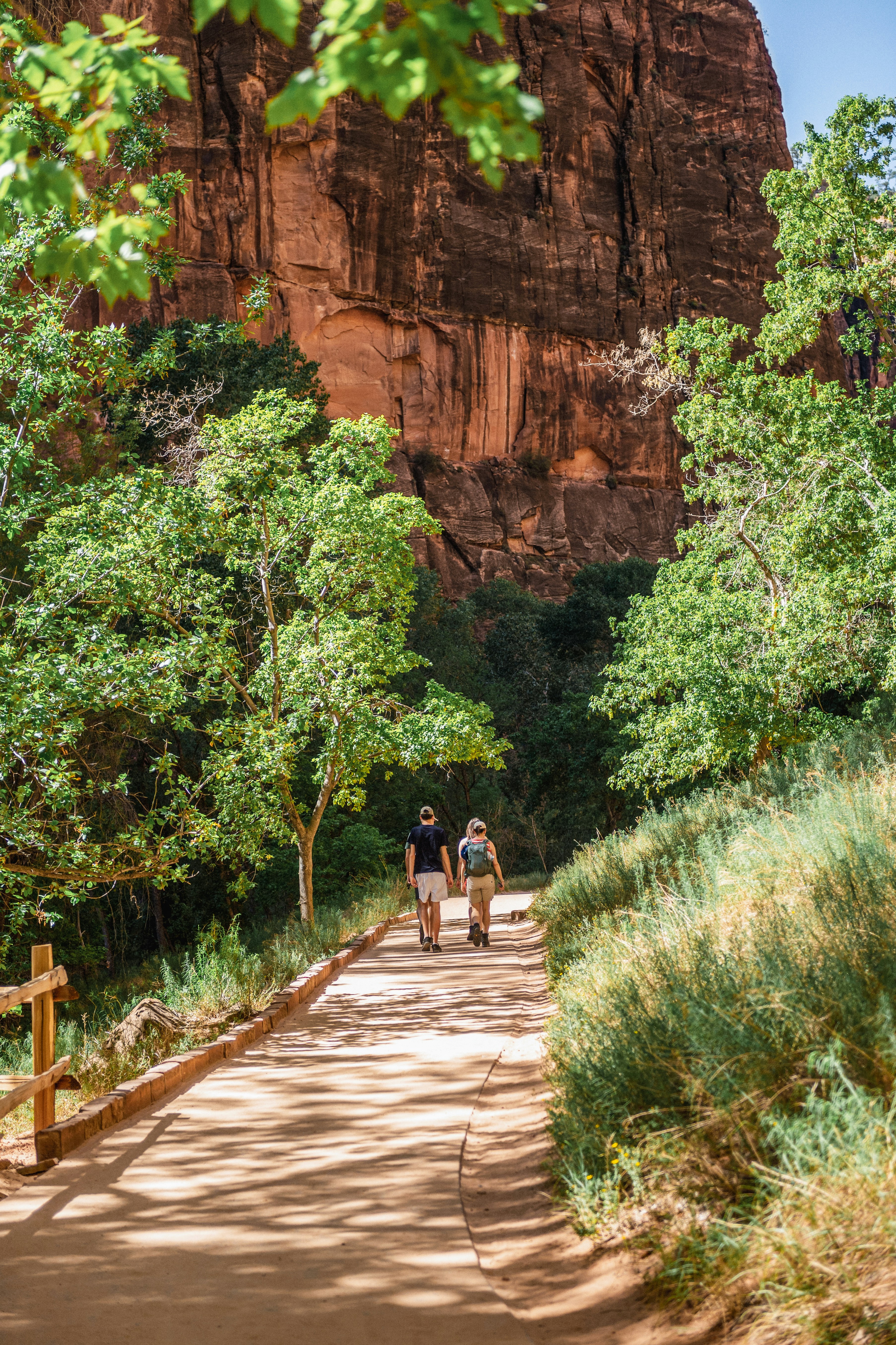 Two hikers walk on a desert trail near red cliffs.