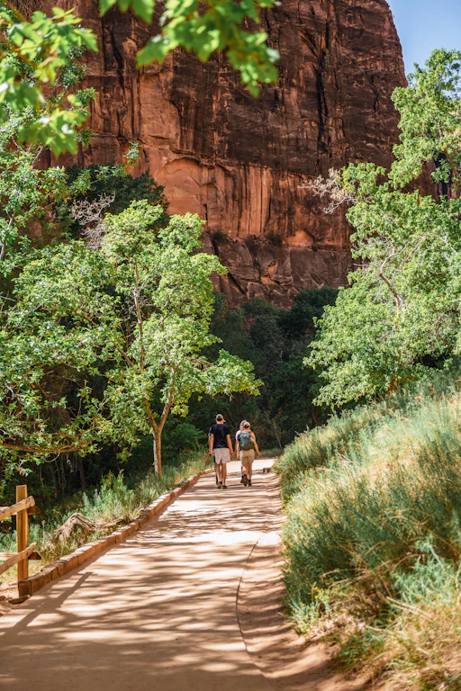 Two hikers walk on a desert trail near red cliffs.