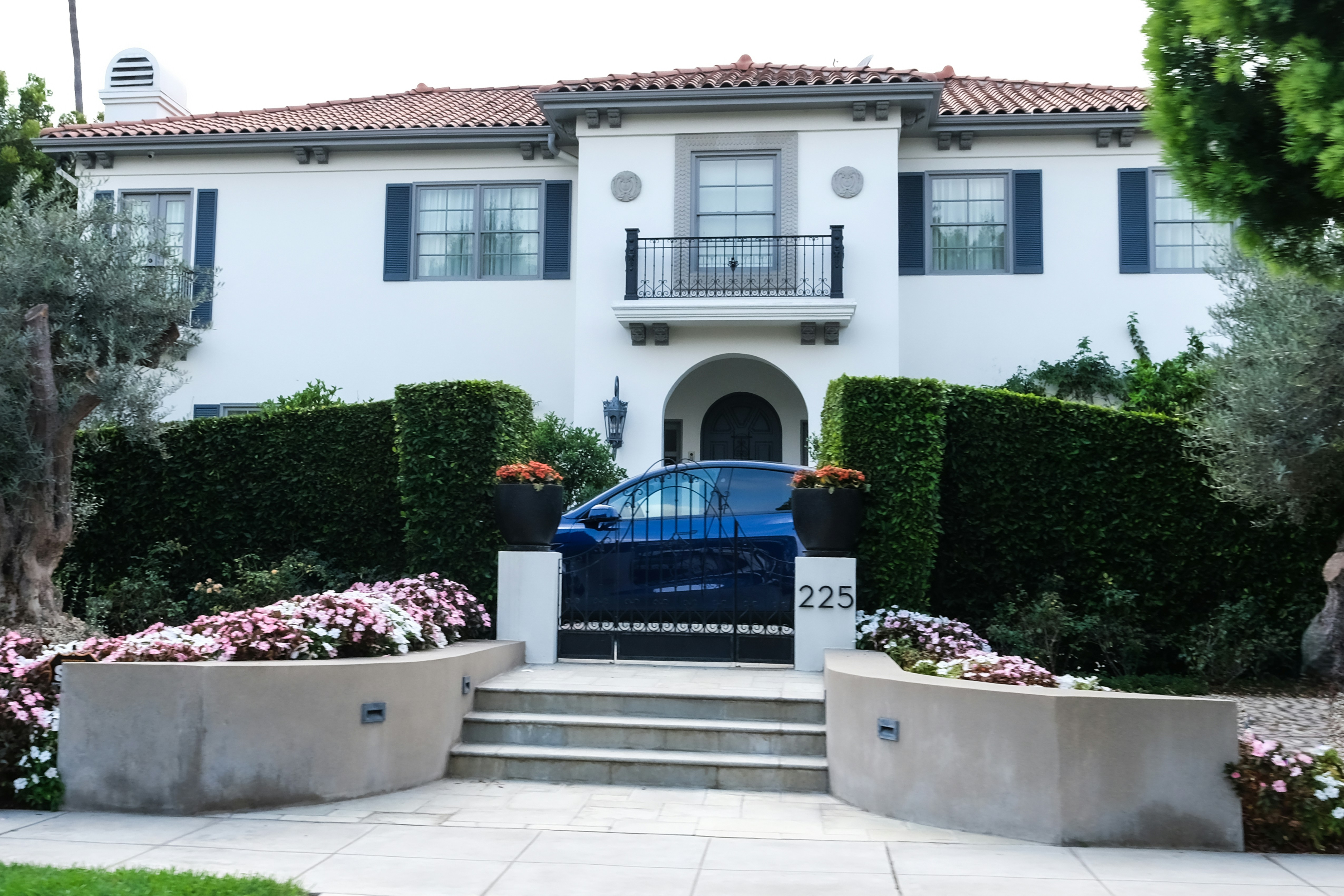 A grand white two-story house with a red-tiled roof and dark blue shutters stands behind a tall hedge. In front of the hedge, a short gate with the number "225" on a post opens to a driveway where a dark blue car is parked