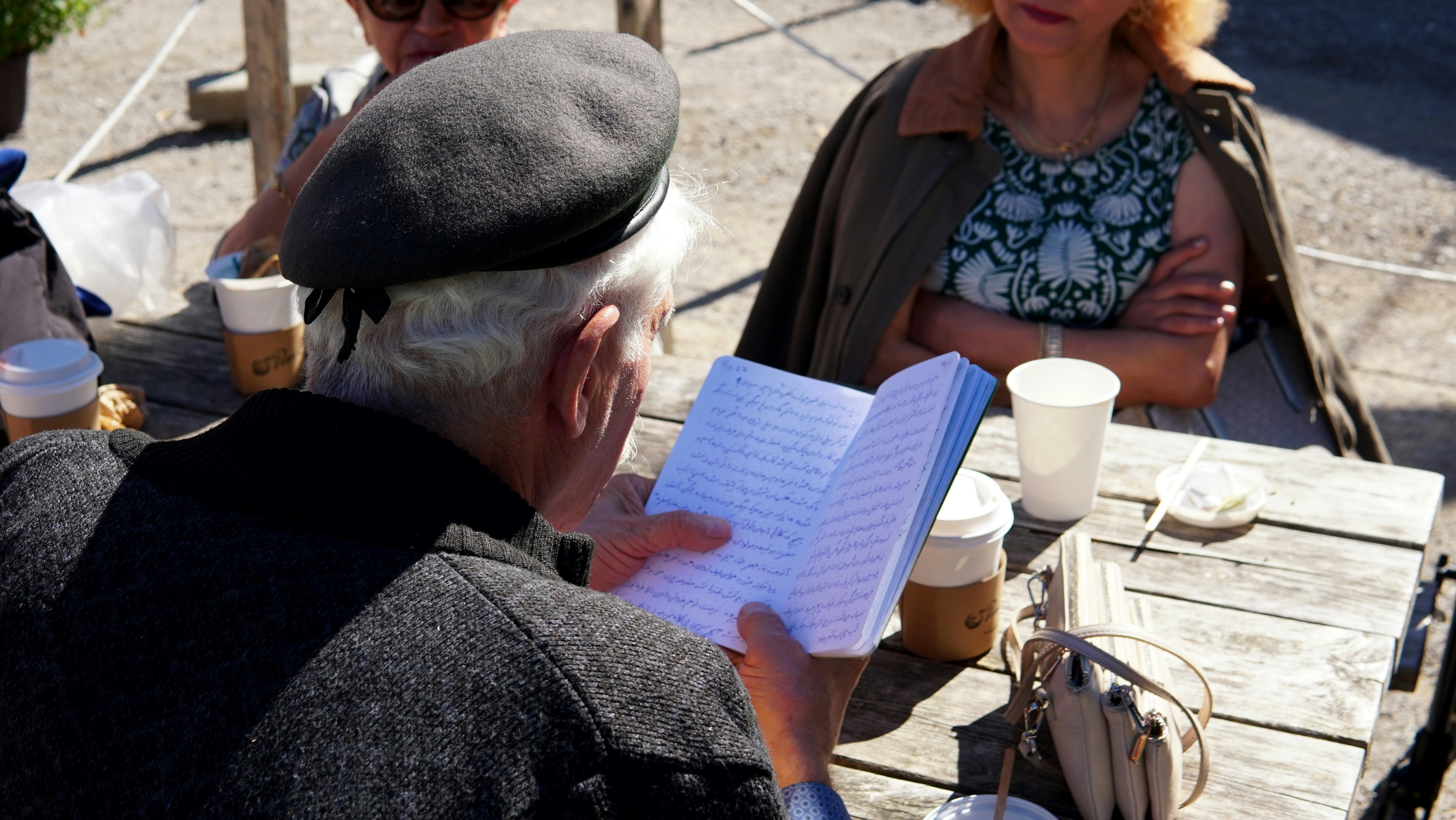 Hombre con boina lee un cuaderno en una mesa al aire libre.