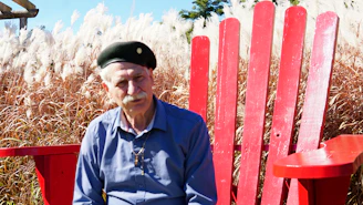 Elderly man in beret sits in large red chair.