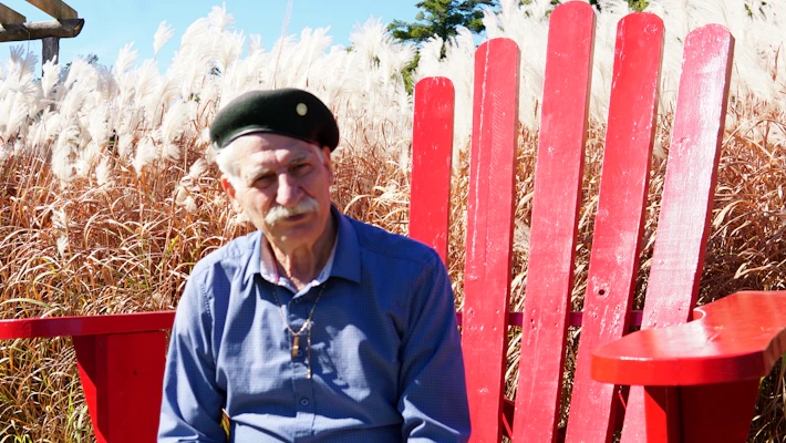 Elderly man in beret sits in large red chair.