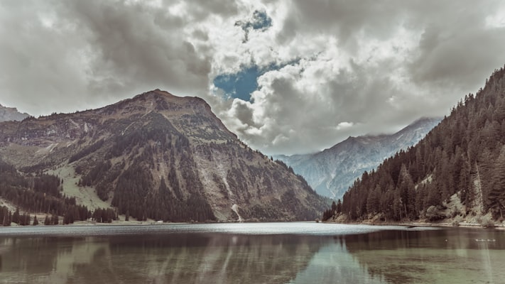 Calm lake reflecting mountains and forest under cloudy sky