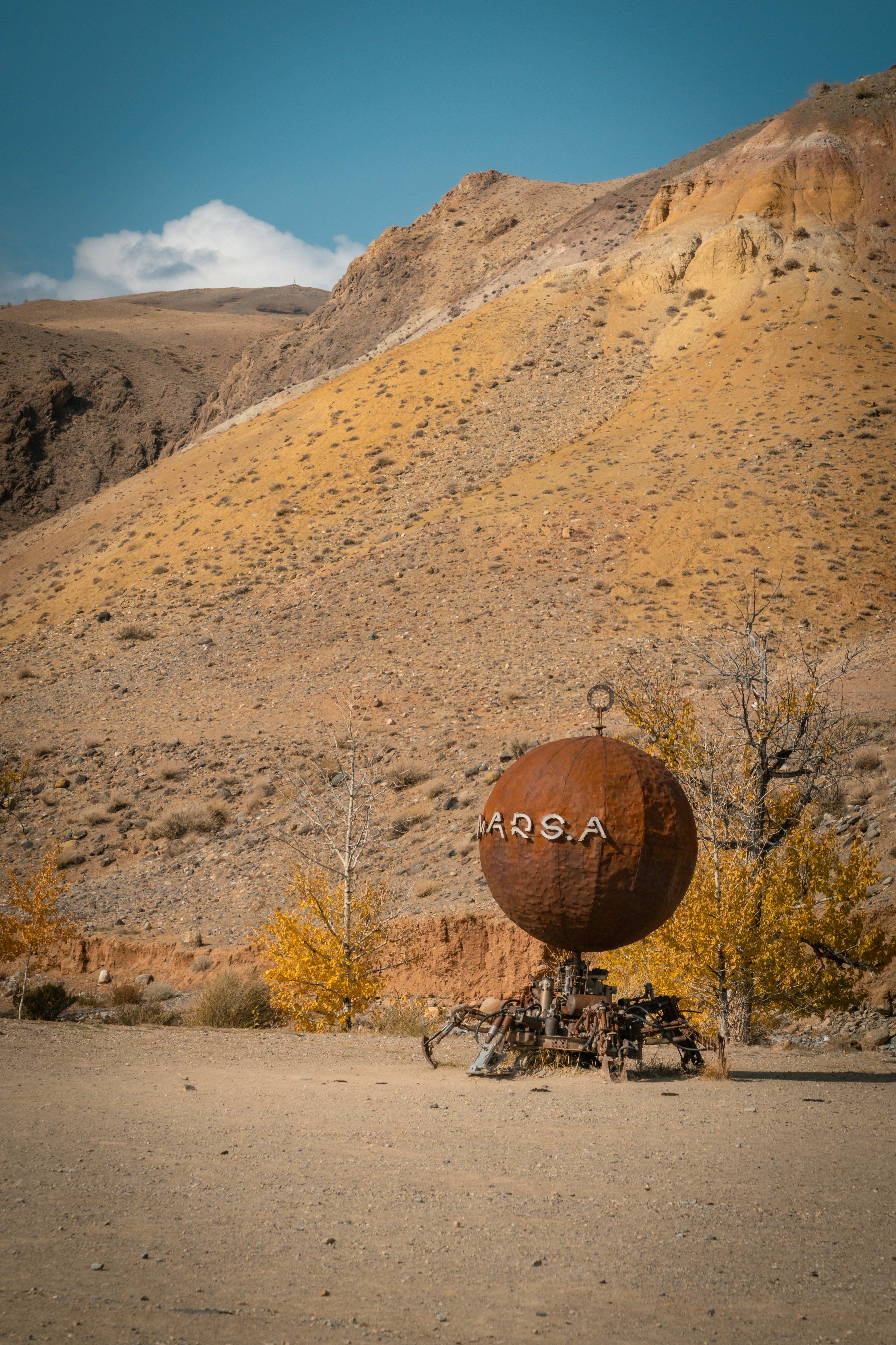 Rusty sphere sculpture in a dry, mountainous landscape.