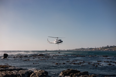 Helicopter hovers over rocky coastline with ocean waves.