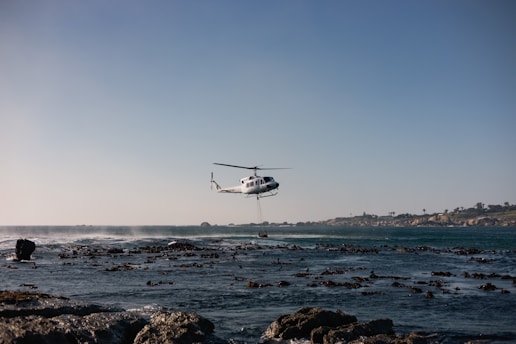 Helicopter hovers over rocky coastline with ocean waves.