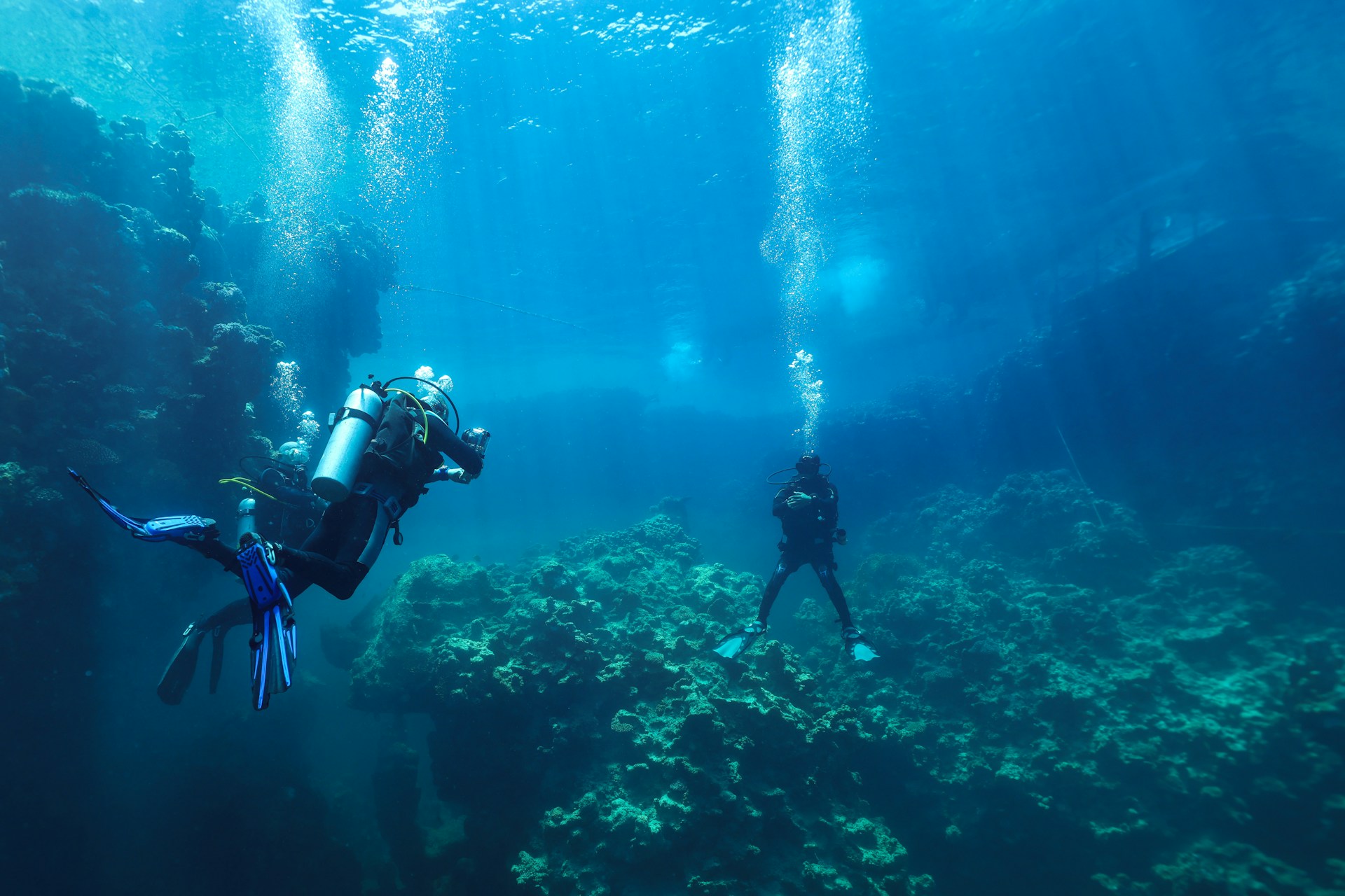 Divers explore a coral reef underwater with bubbles rising.