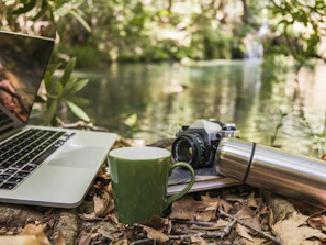Laptop, camera, and mug by a forest stream