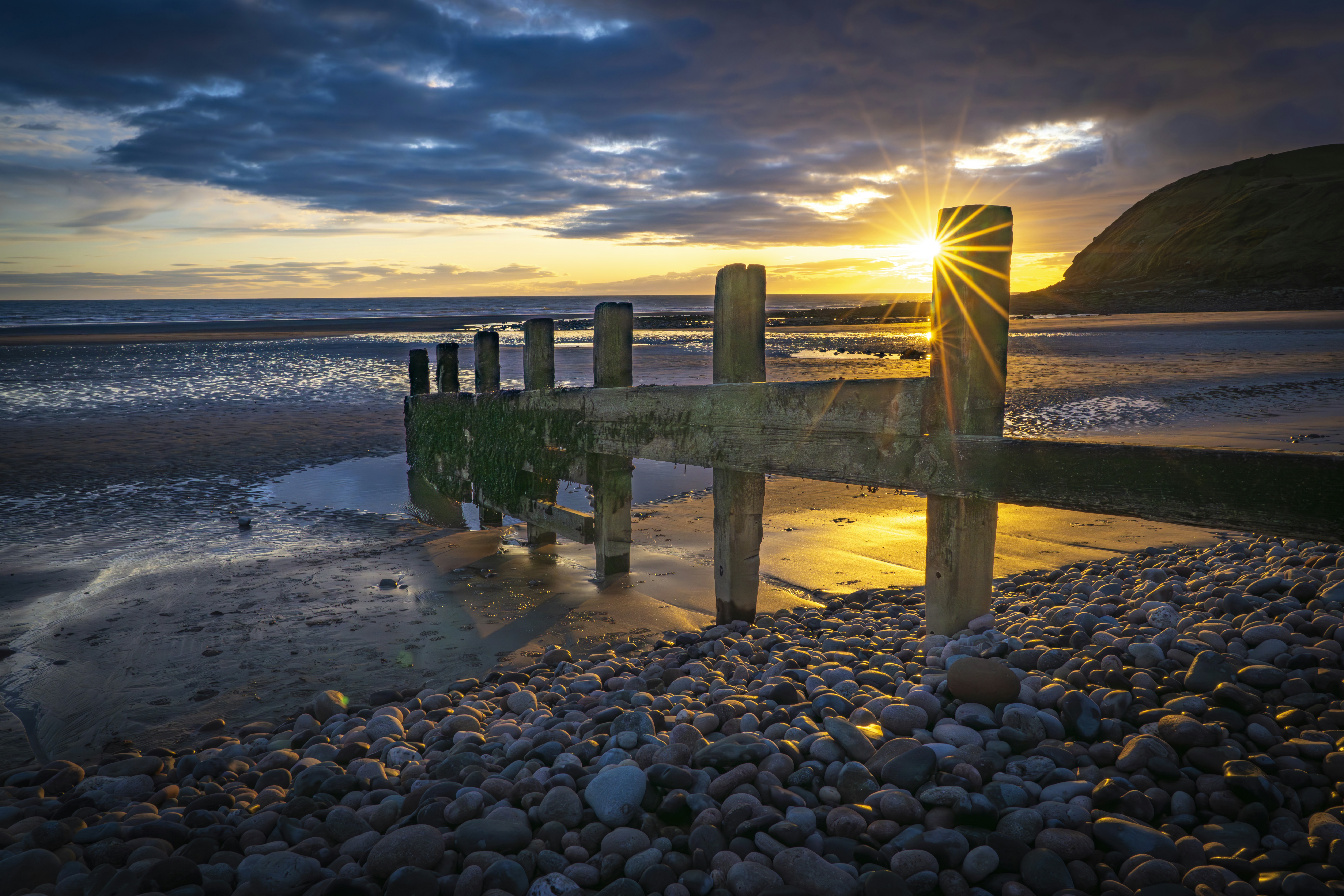 Sunrise over a pebble beach with wooden structure.