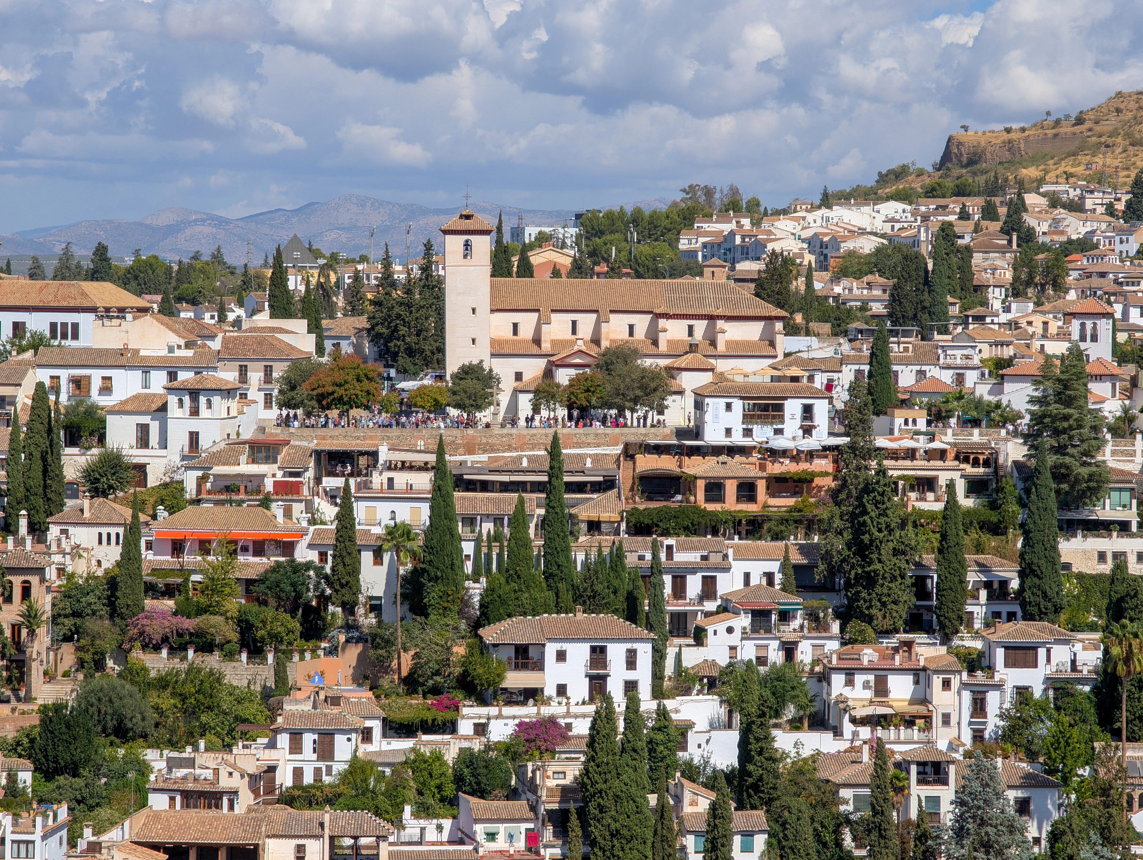 The Albaicin quarter of Granada climbing its hillside in whitewashed houses and terracotta roofs among dark cypresses, with the stone tower of San Nicolas church on the ridge and mountains and piled cloud behind.