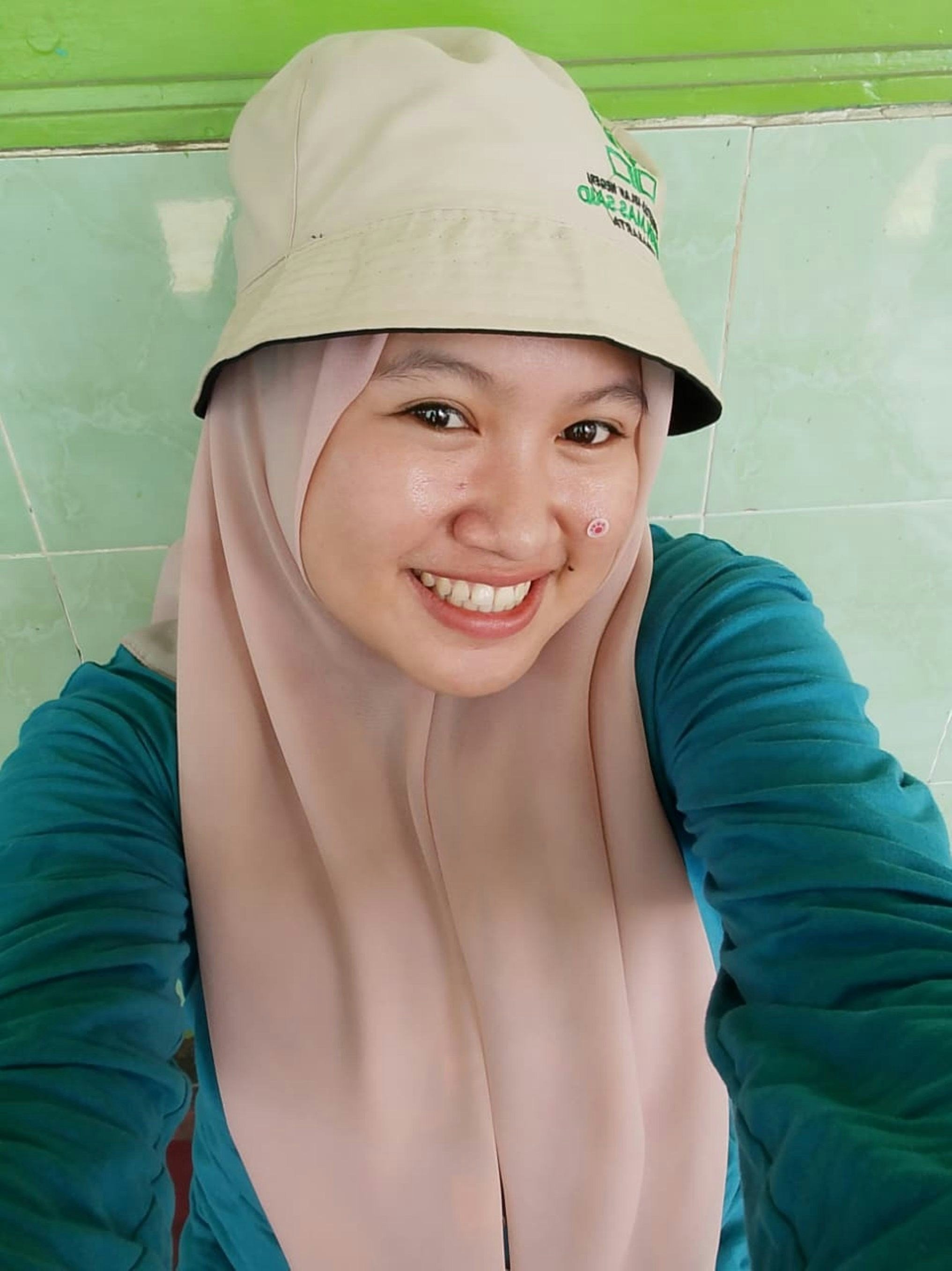 Seorang wanita muda yang mengenakan topi ember dan jilbab tersenyum.