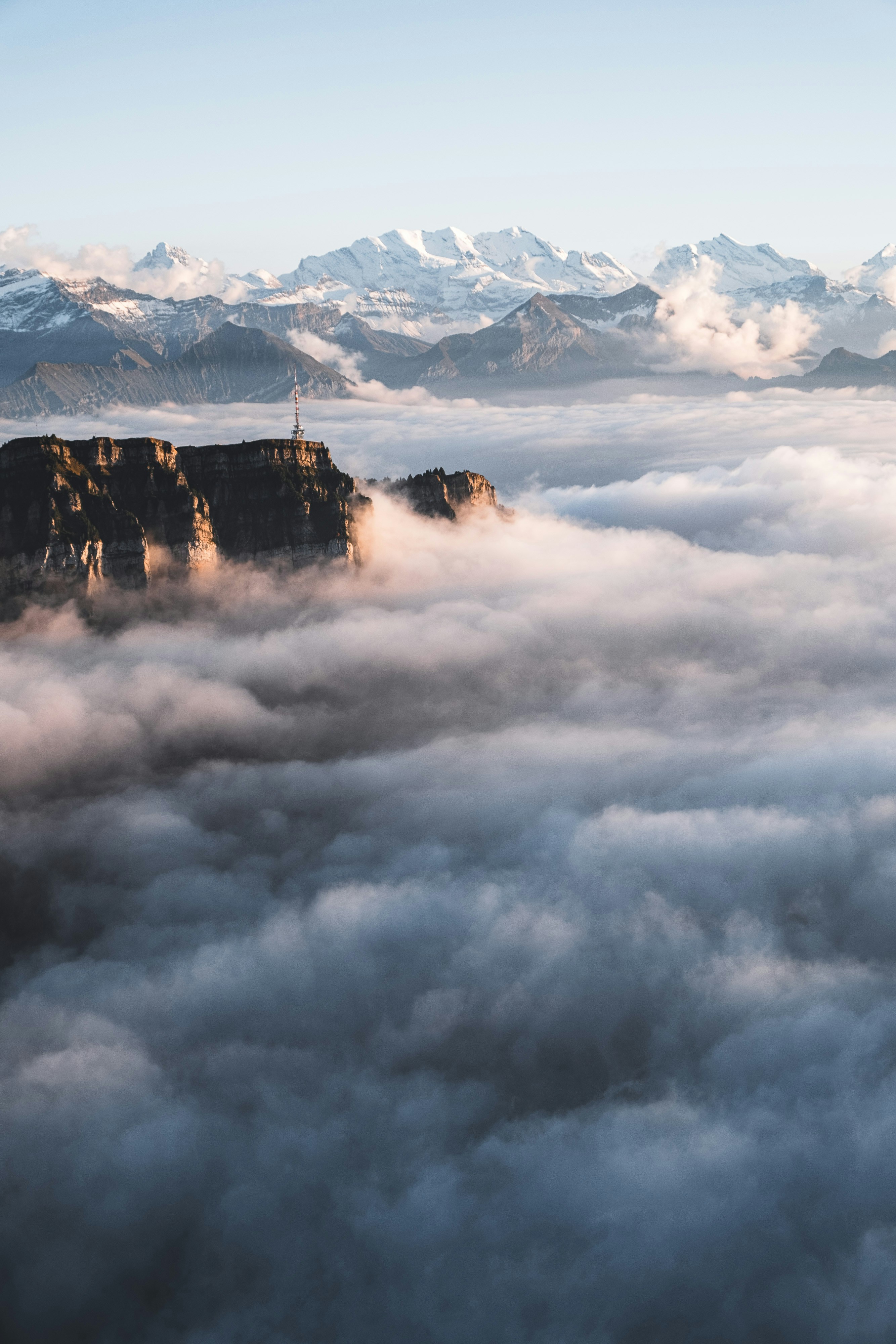 Rocky cliff above clouds with snowy mountains beyond.