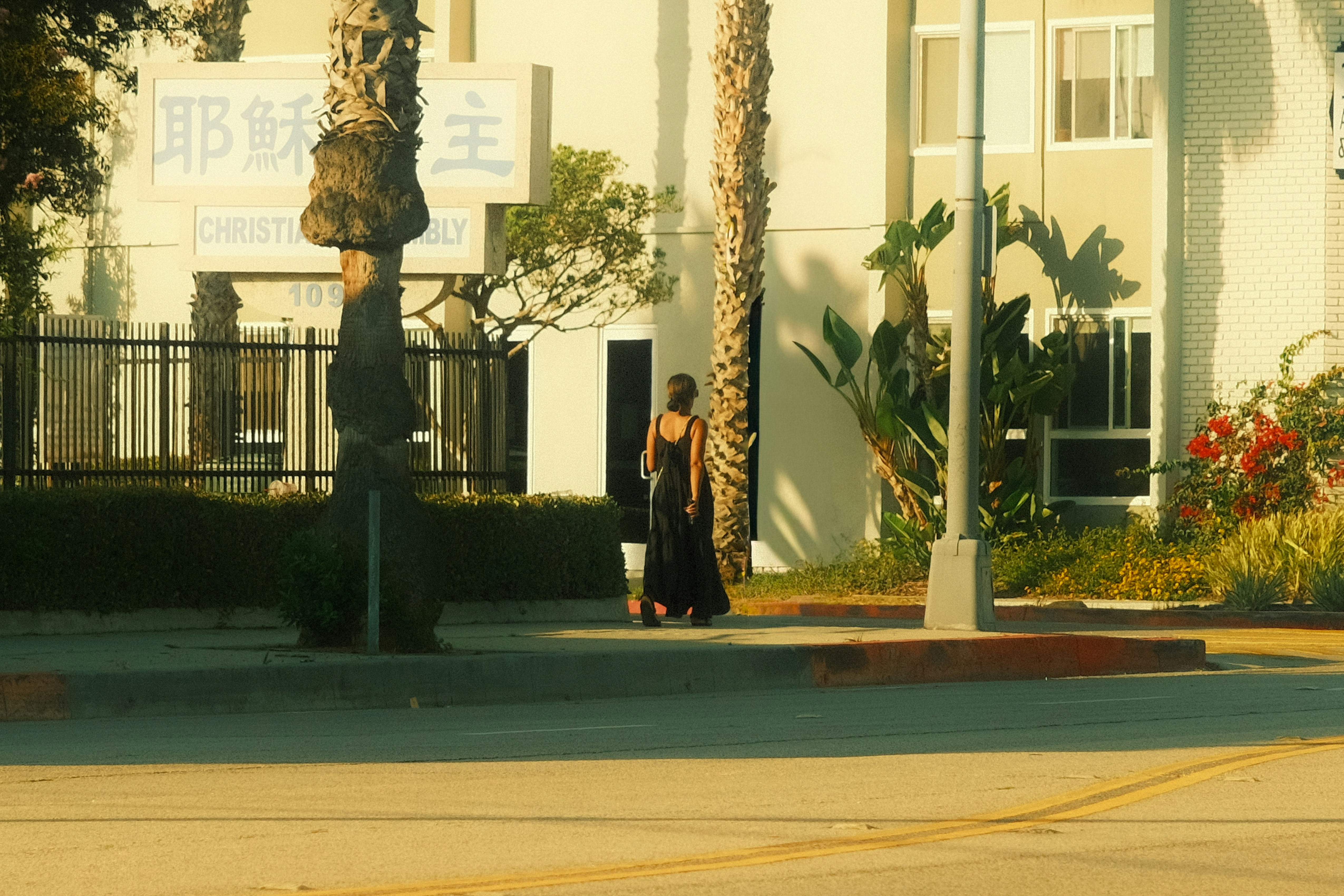 Woman in black dress walks on sidewalk near building.