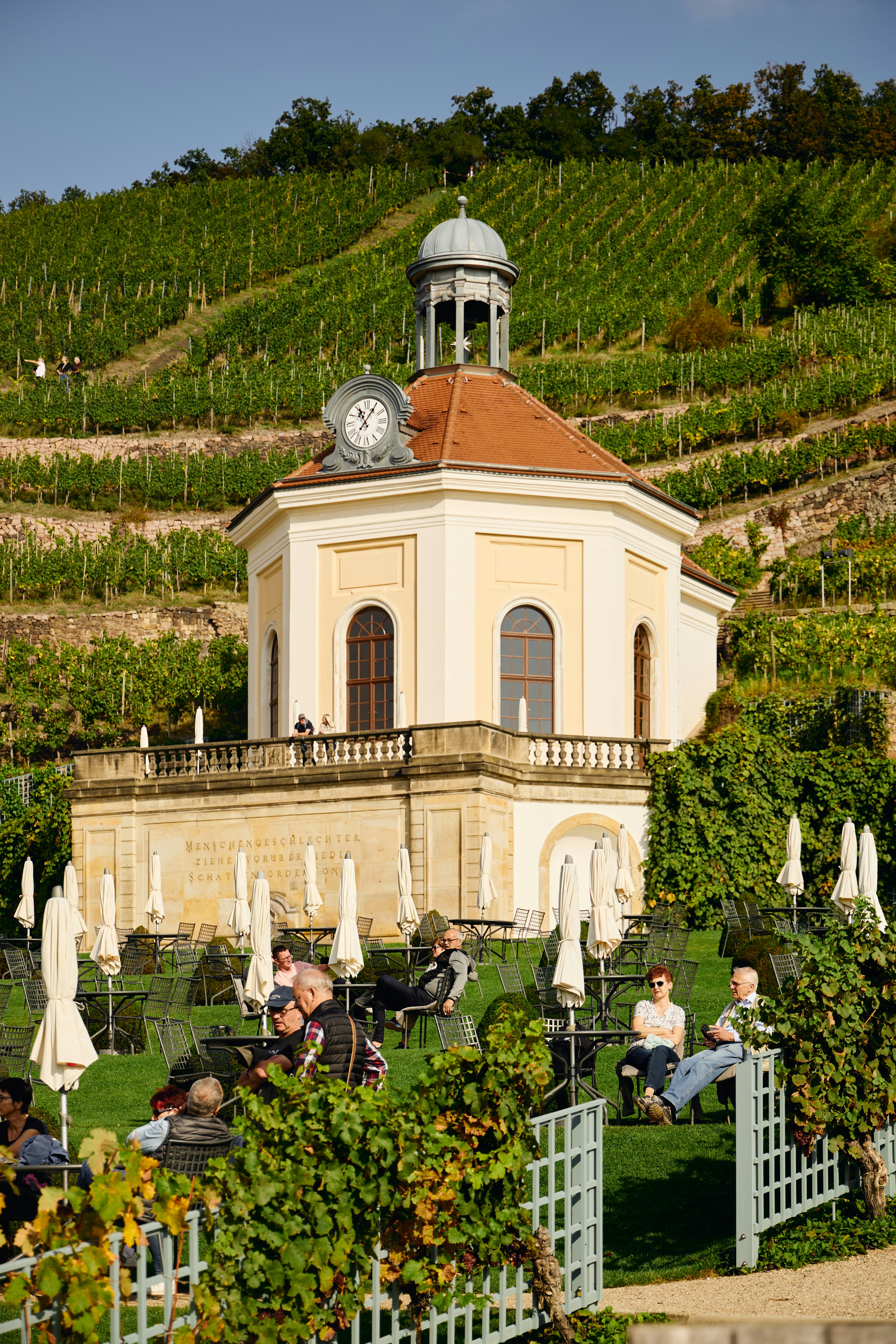 People relax at tables near a building in a vineyard.