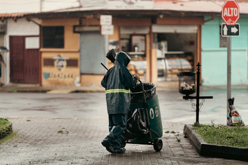 Sanitation worker pushing trash cart on wet street.