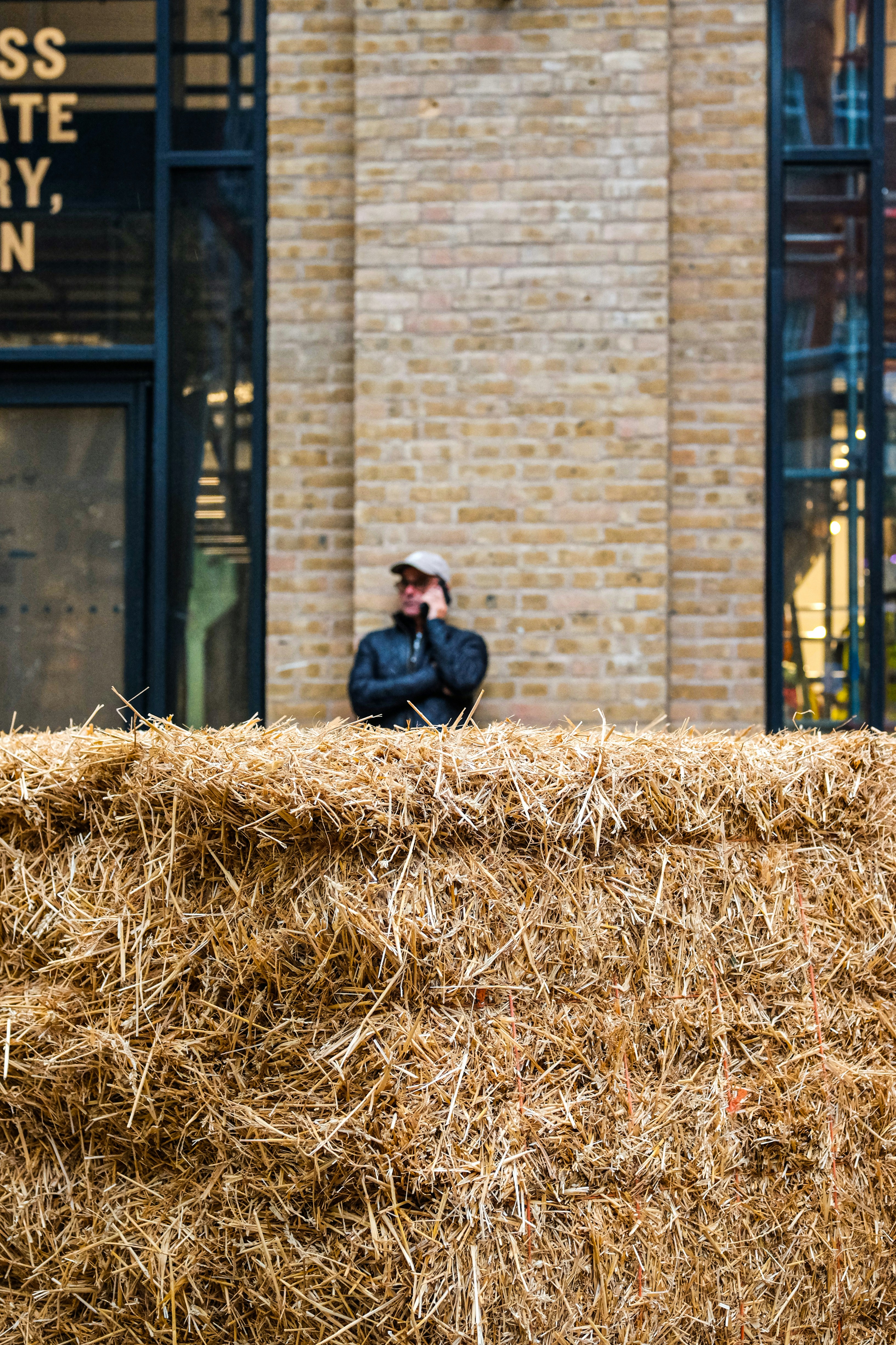 Man in hat stands behind hay bales talking on phone.