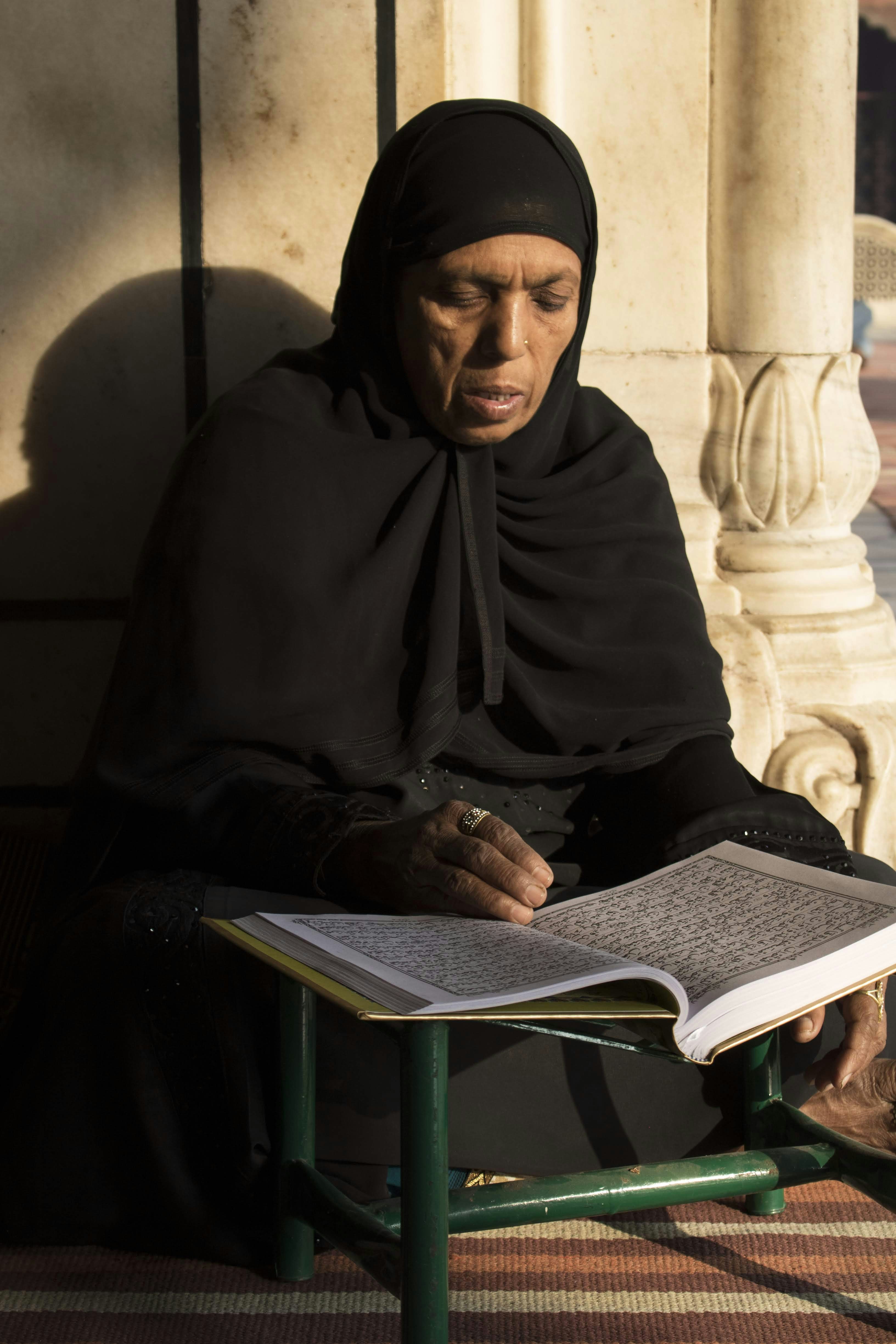 Elderly woman in hijab reads from an open book.