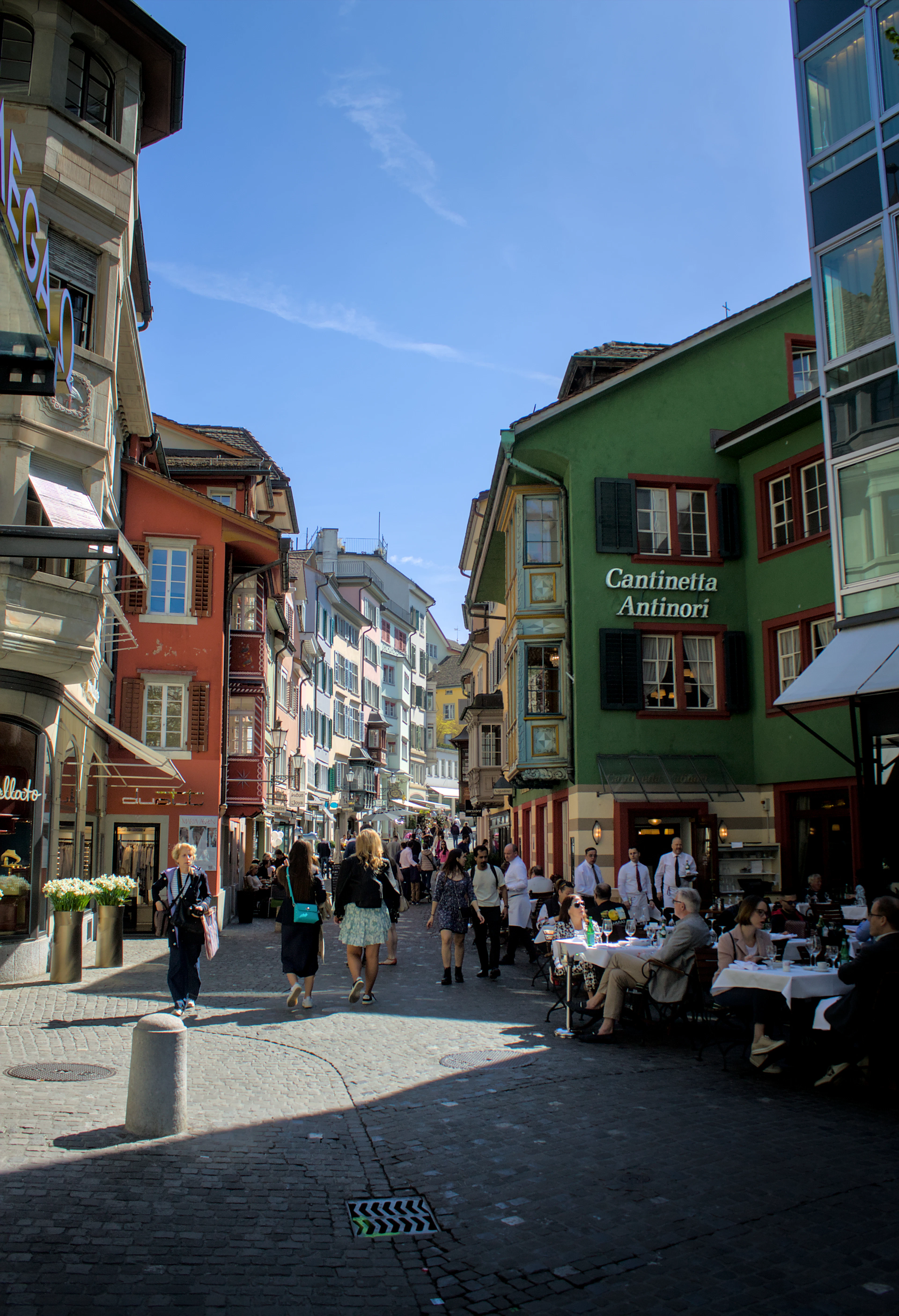 Colorful painted facades line a sunlit old-town street with cafe tables and strolling pedestrians.