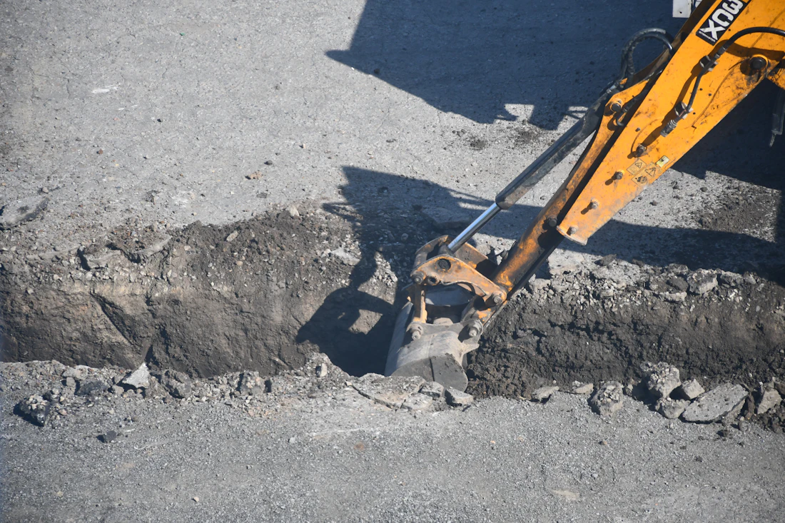Excavator bucket working in an open utility trench cut through asphalt pavement