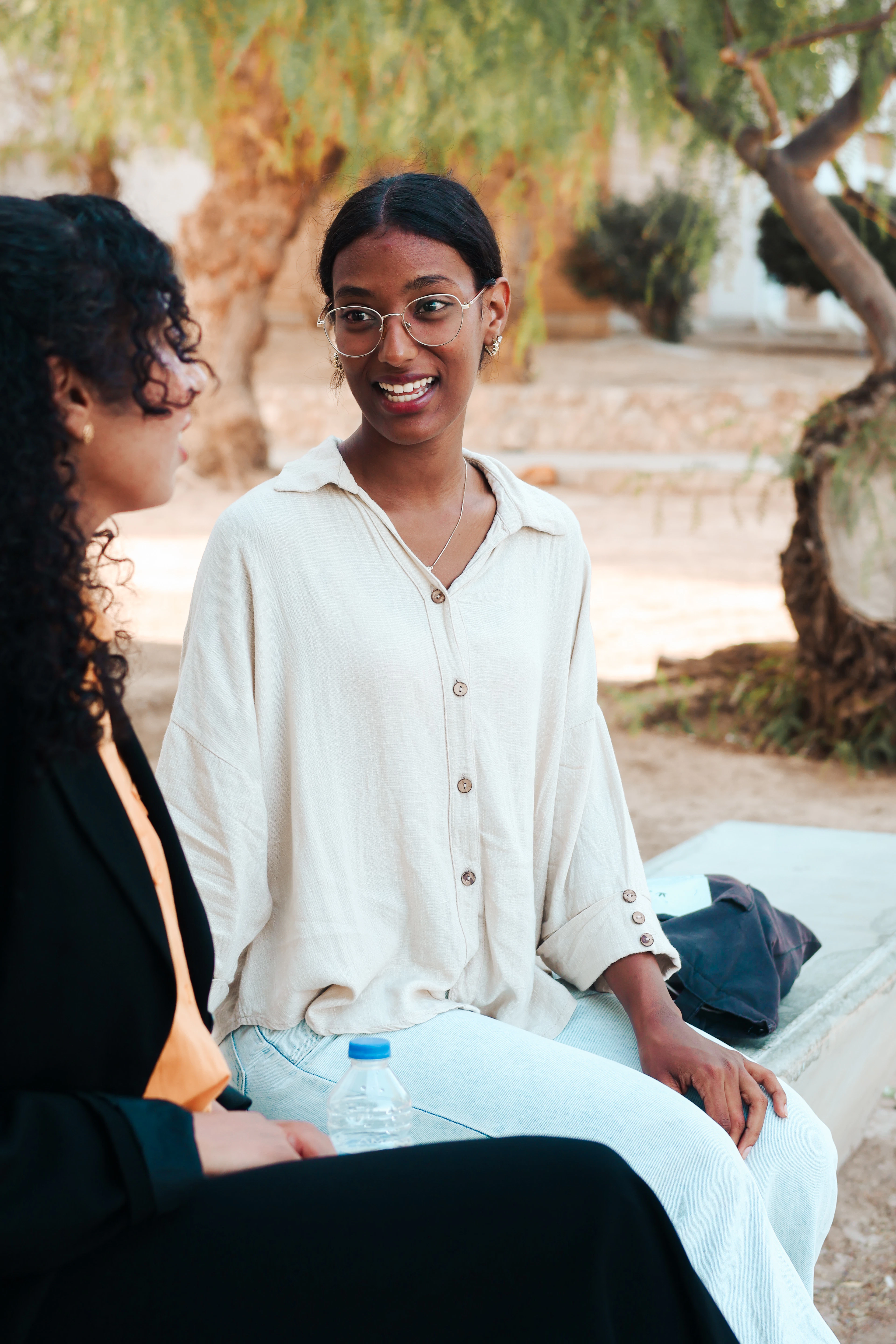 Two women in conversation outdoors under warm afternoon light, one wearing glasses and smiling