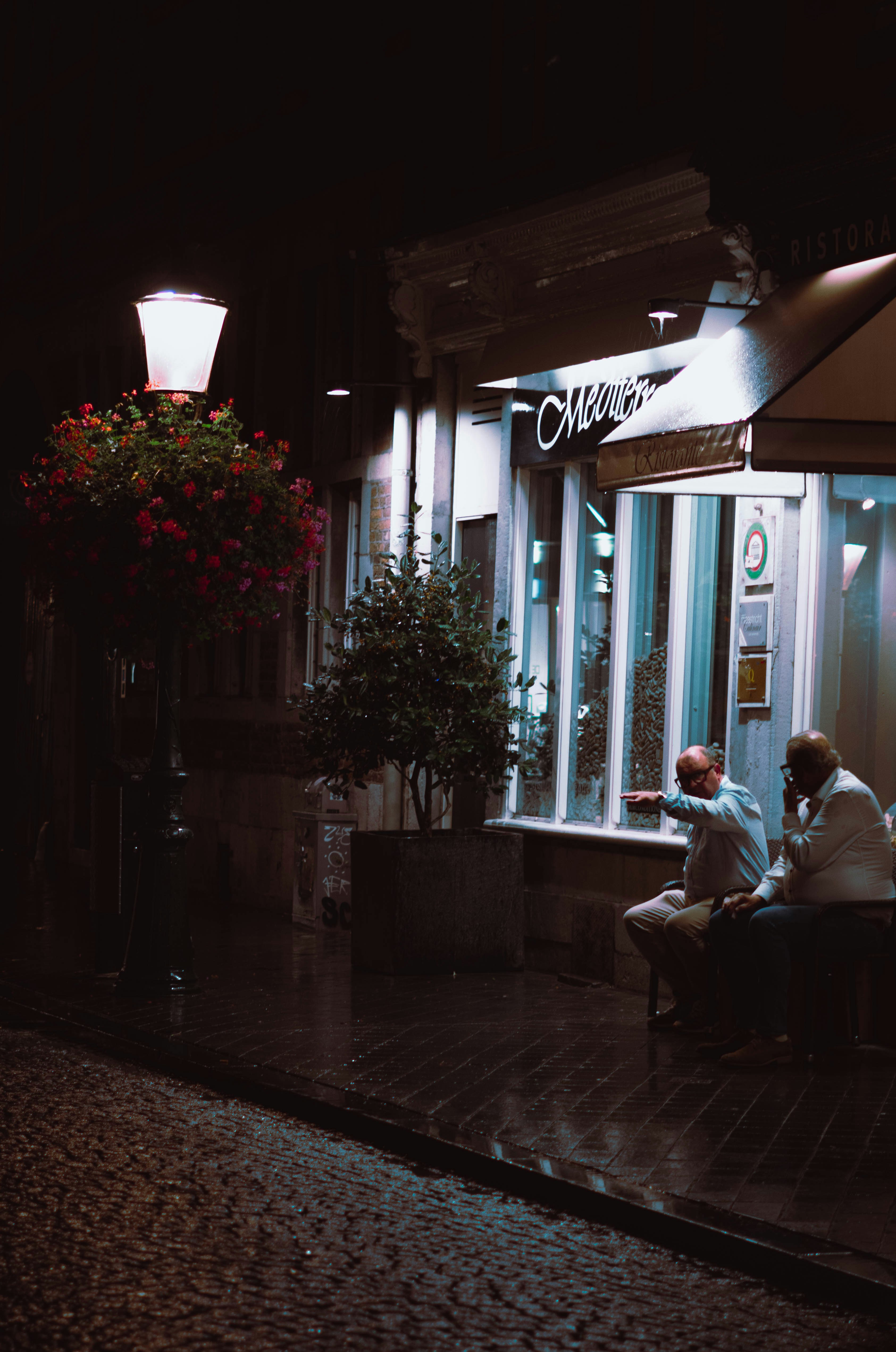 Dos hombres se sientan afuera de un café bien iluminado por la noche.