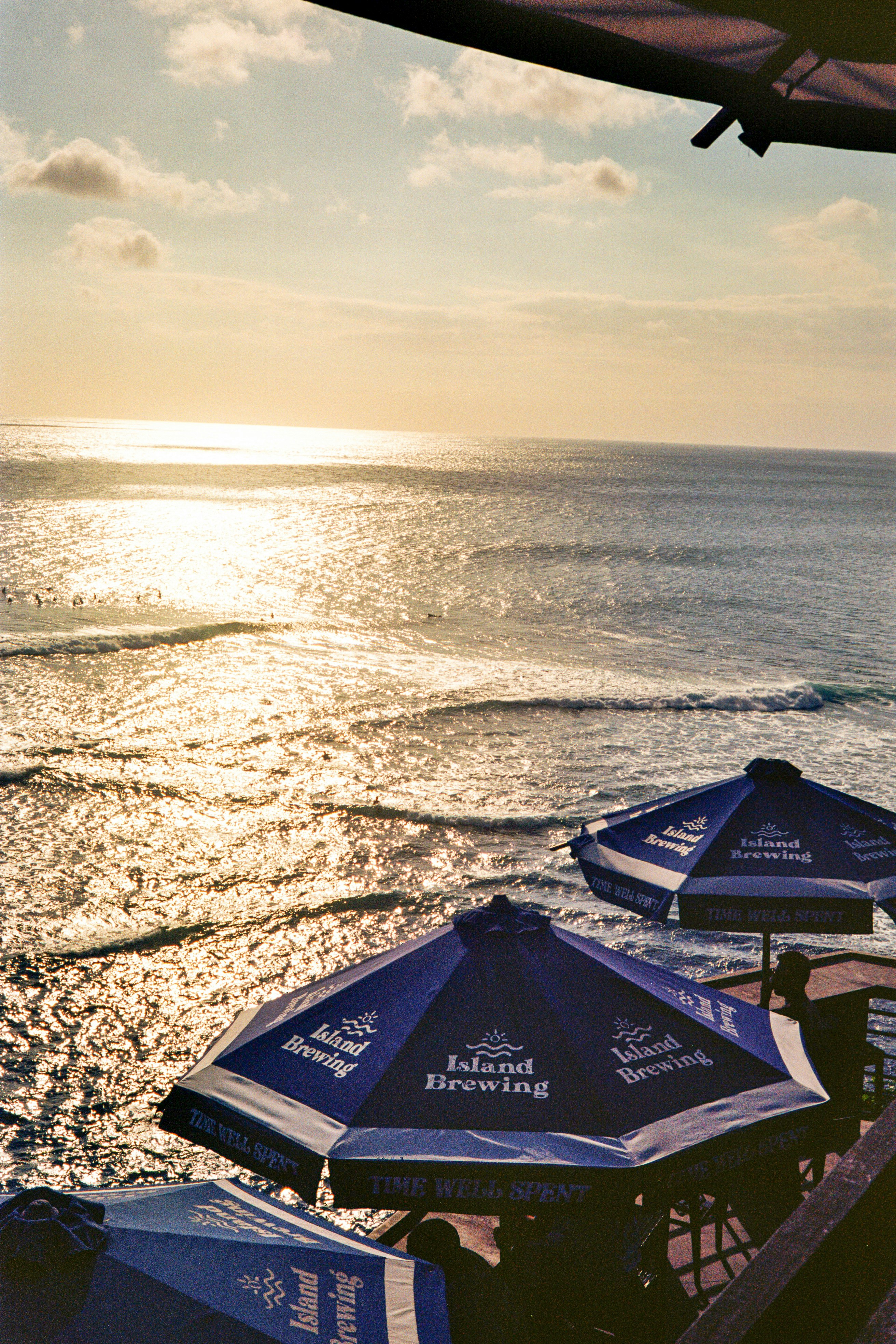 Beachside umbrellas from Island Brewing overlook tranquil ocean waves reflecting the soft glow of the setting sun.